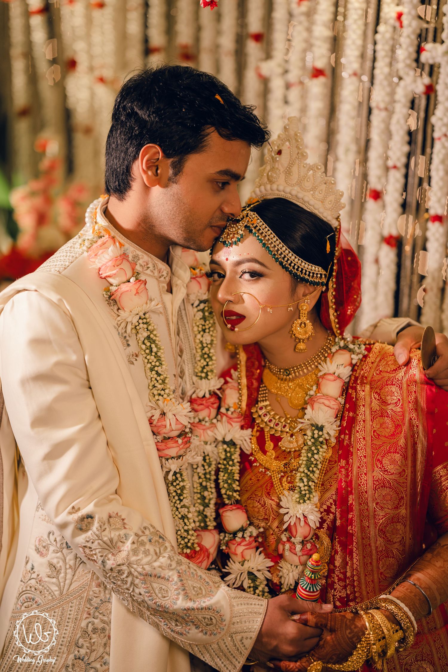Wedding couple in traditional Indian attire sharing intimate moments against floral backdrop.