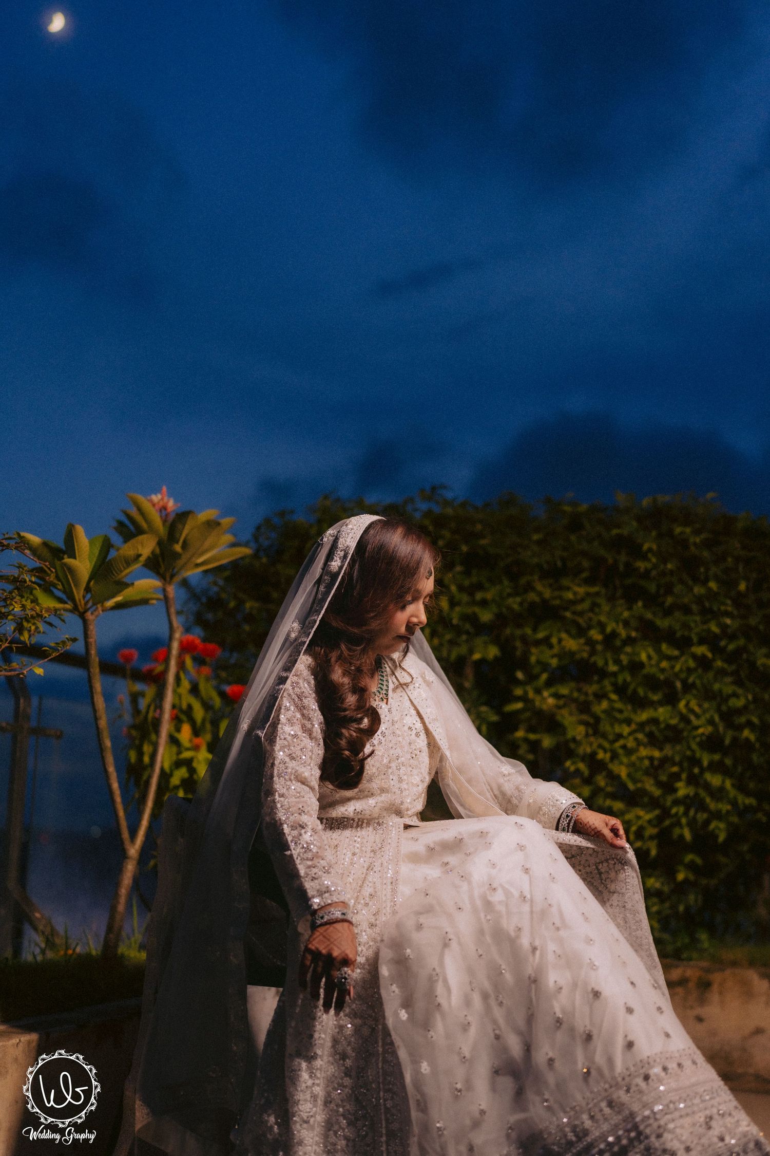 Bride in white lace gown and veil sits outdoors at dusk with palm trees and moon in background.