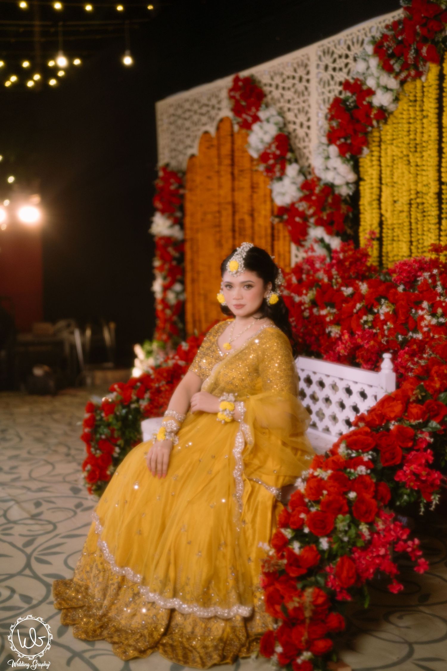 Beautiful yellow lehenga set against a backdrop of red flowers and white lattice decor with string lights overhead.