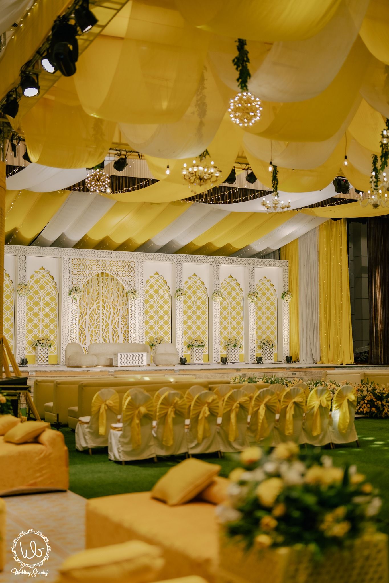Elegant wedding venue with draped yellow fabric ceiling, ornate white stage backdrop, and crystal chandeliers.