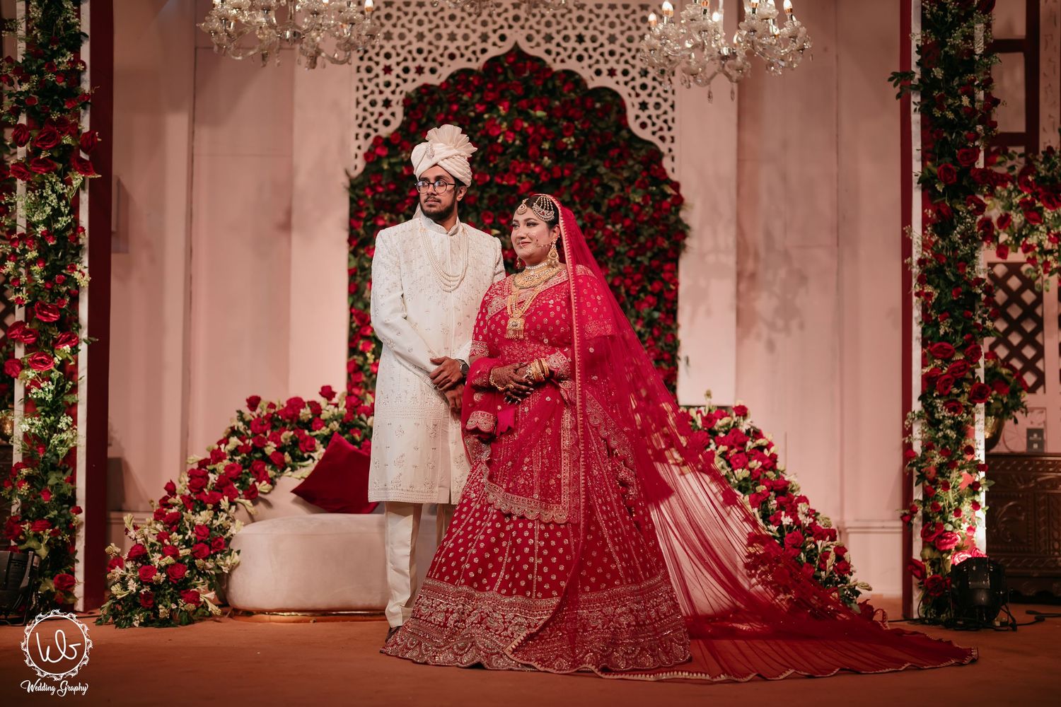 Couple in traditional Indian wedding attire poses under floral archway with red roses and soft lighting.