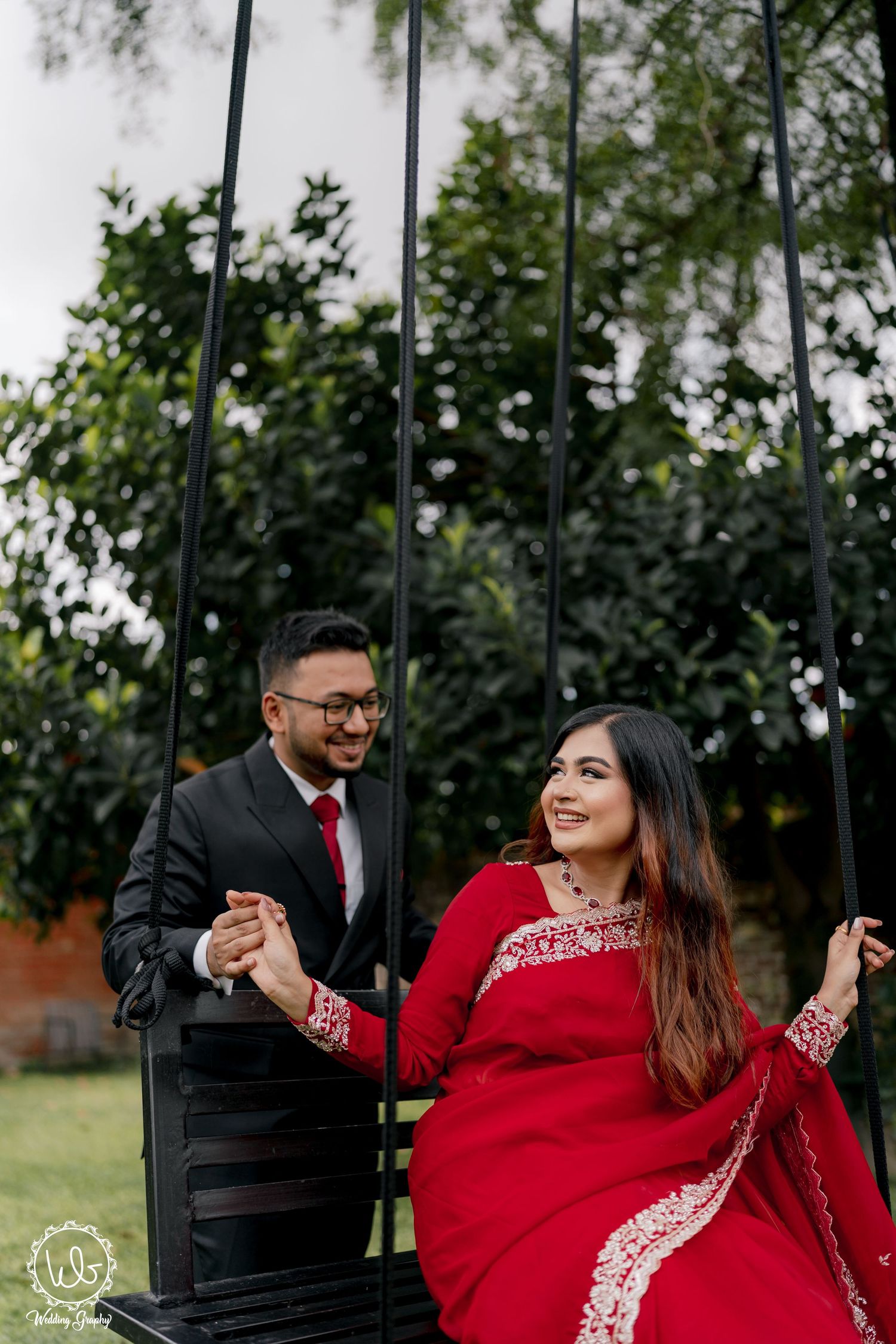 Couple on swing in garden, wearing red and black formal attire.