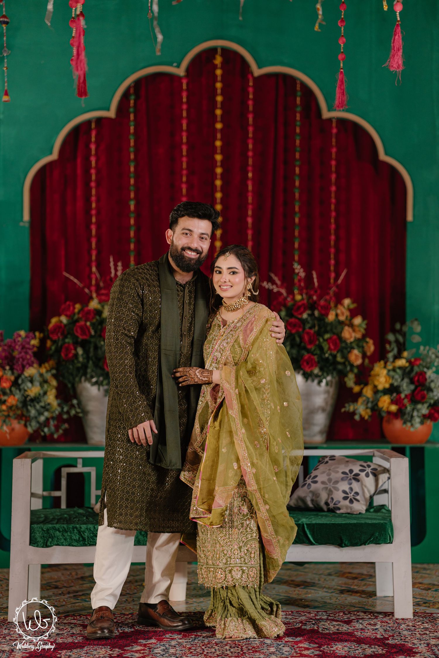 Couple in traditional Pakistani attire poses together at decorated wedding venue with green walls and red drapes.