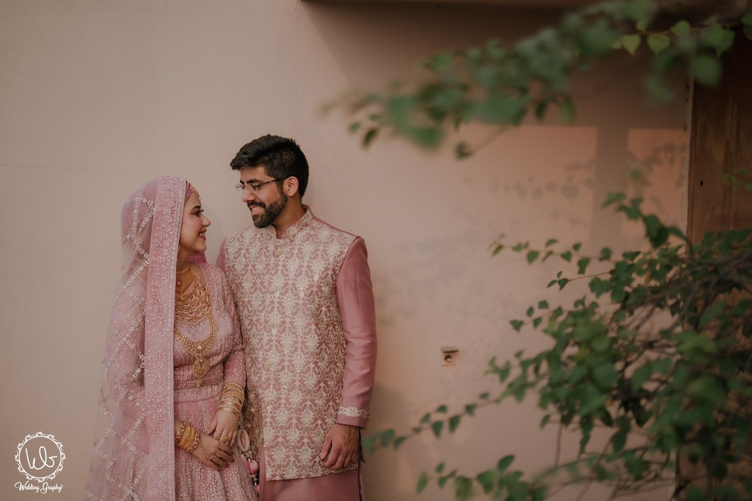 A couple in traditional Indian wedding attire poses together against a soft pink wall with green foliage.