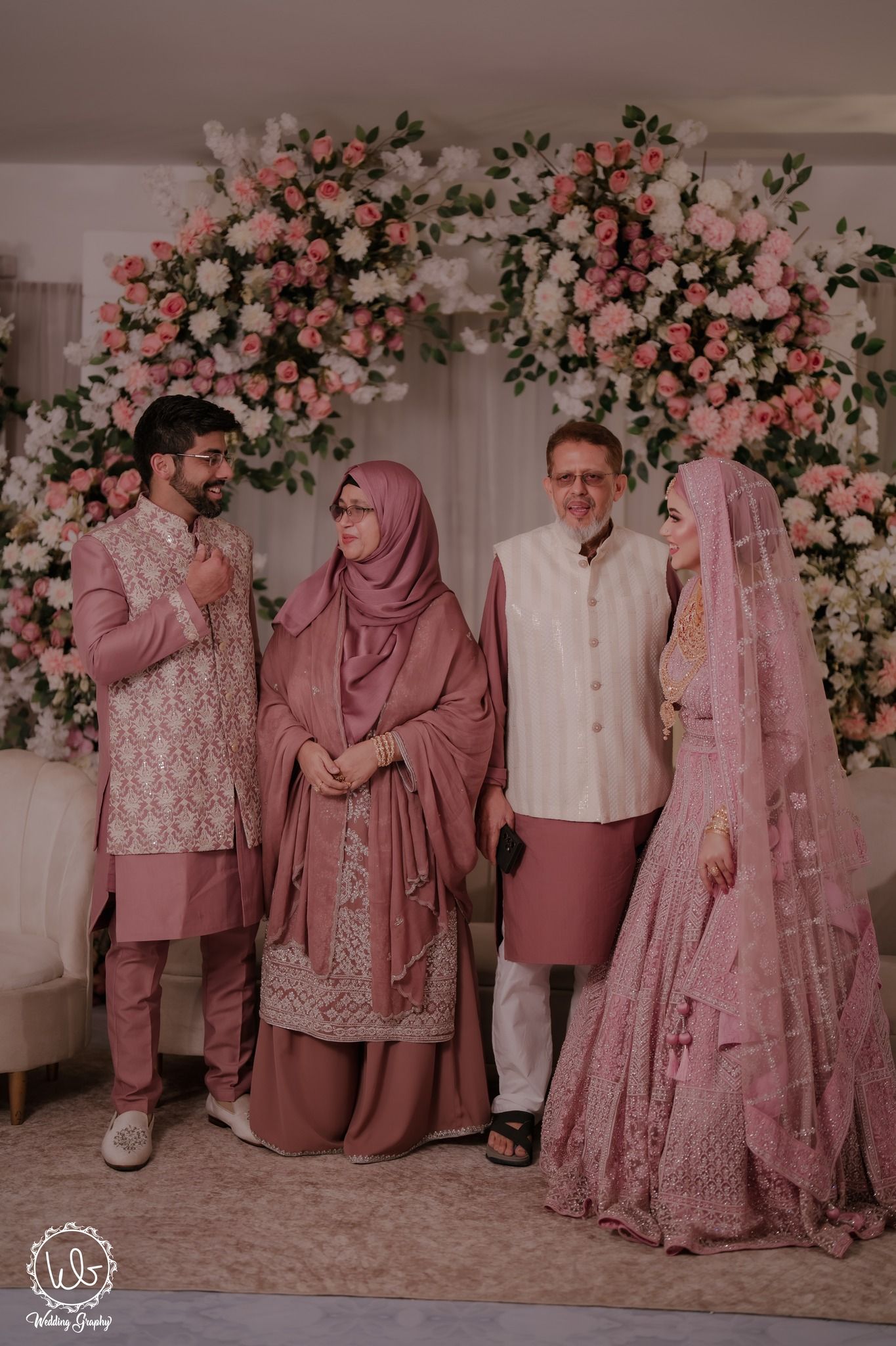 Family wearing coordinated pink and white traditional outfits poses together against floral backdrop for special occasion photos.