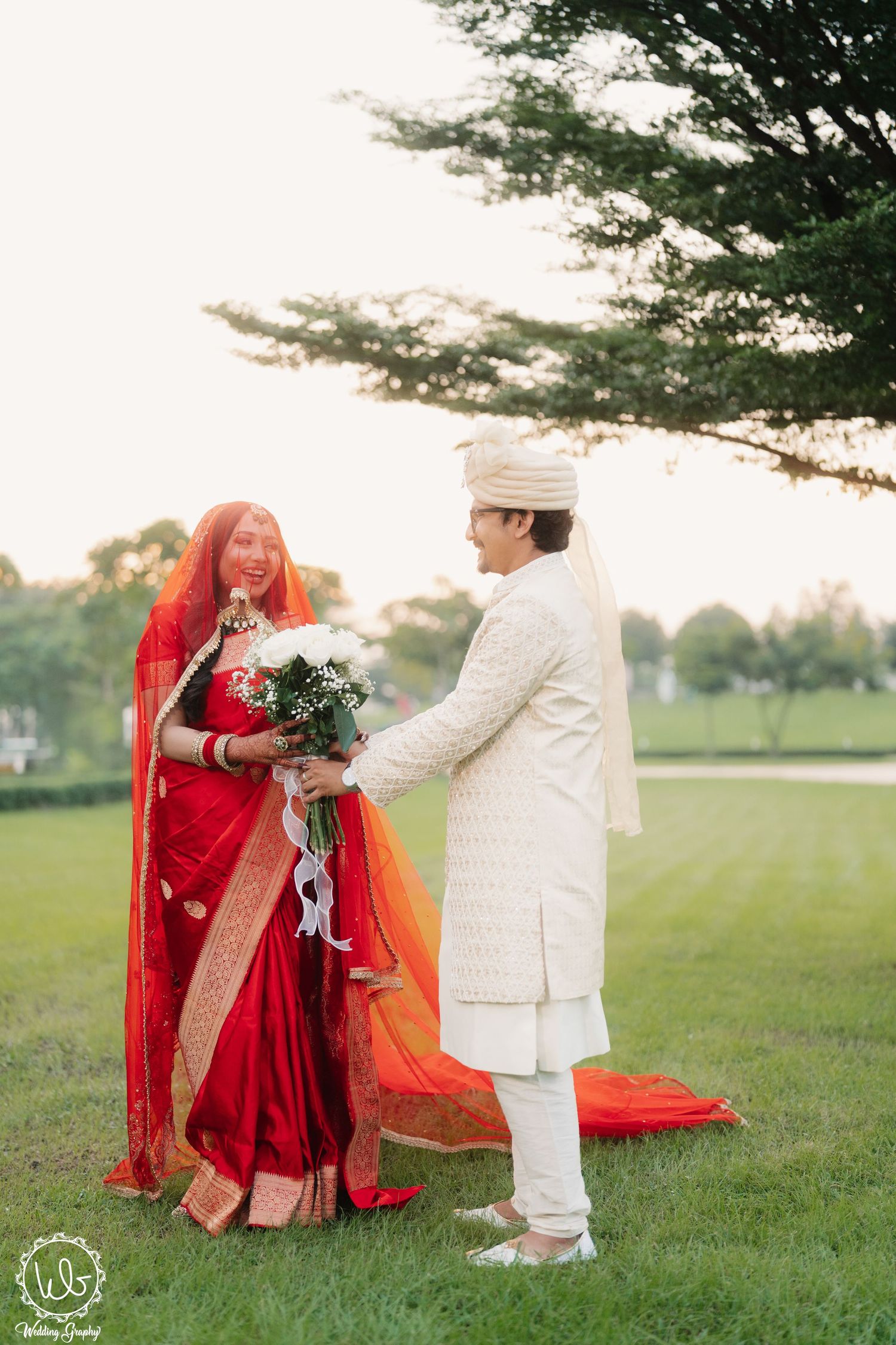 A couple in traditional wedding attire share a romantic moment on a grassy field during sunset.