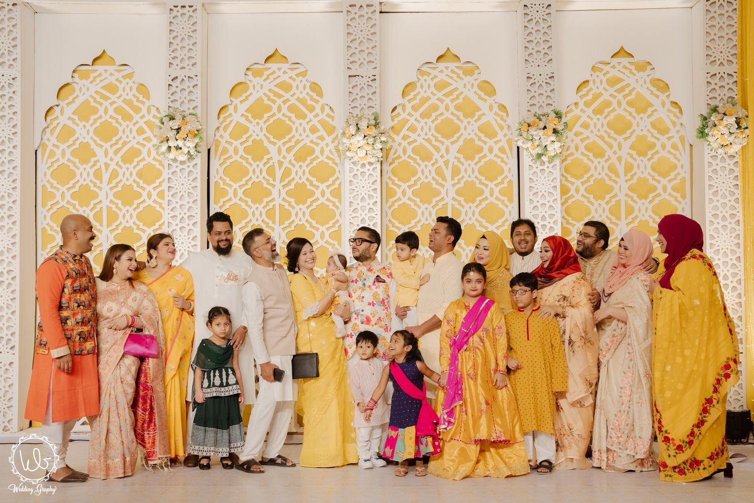 Group photo of wedding guests in traditional attire against an ornate gold and white geometric backdrop.