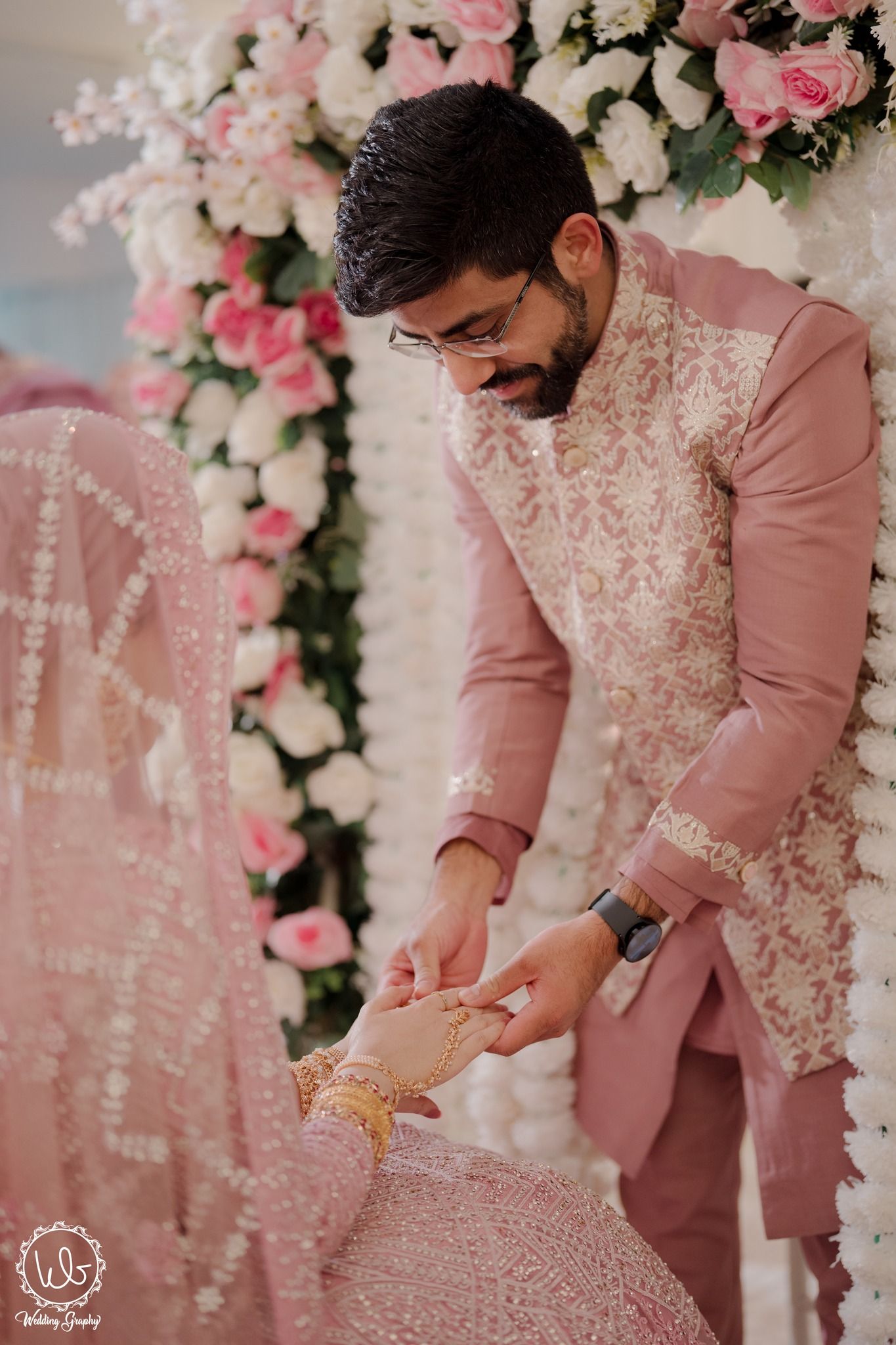 Traditional wedding ceremony with pink floral decor shows a moment of ring exchange against a backdrop of roses.