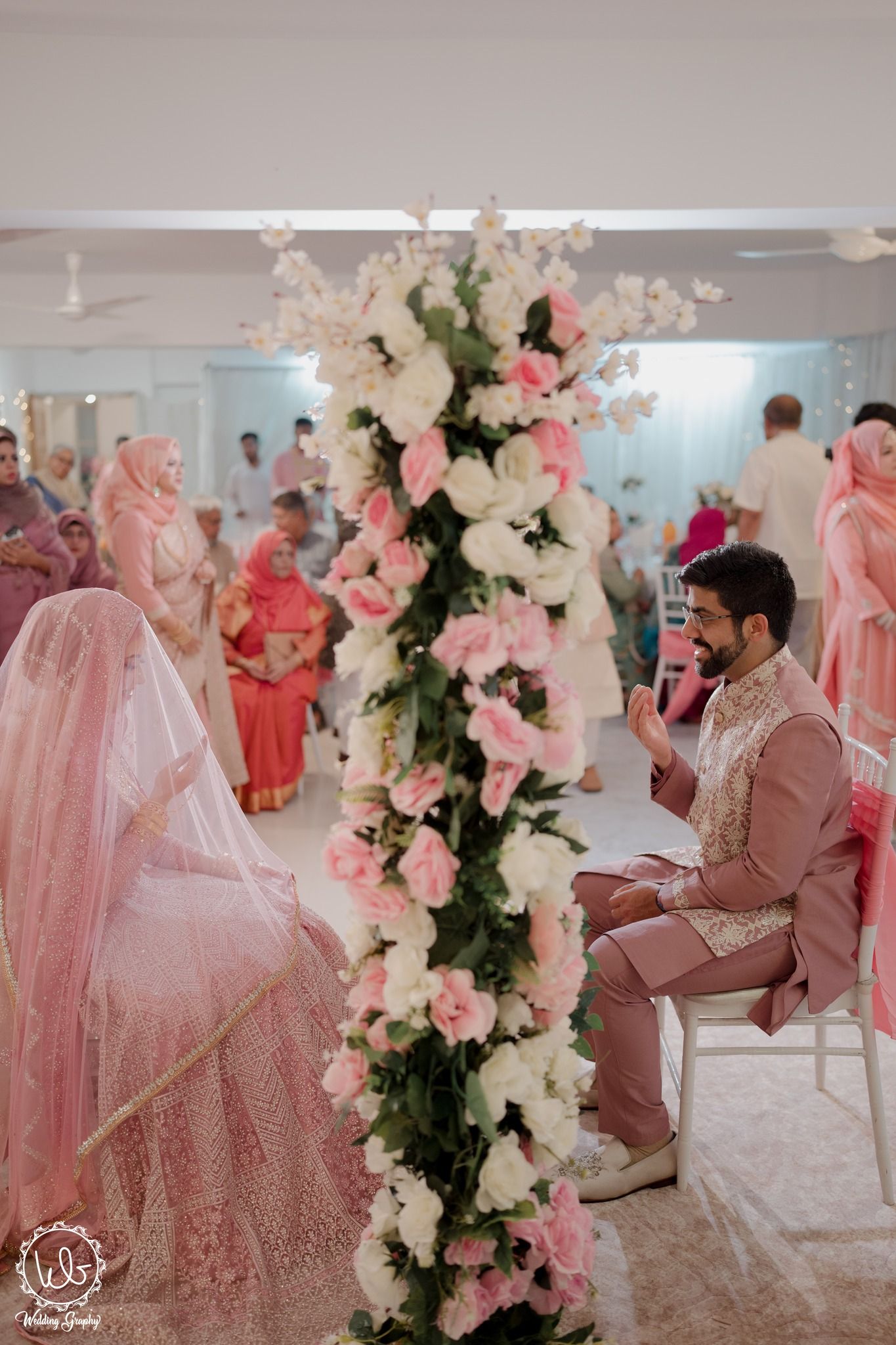 Pink and white floral pillar decorates an elegant wedding ceremony with guests in traditional attire.