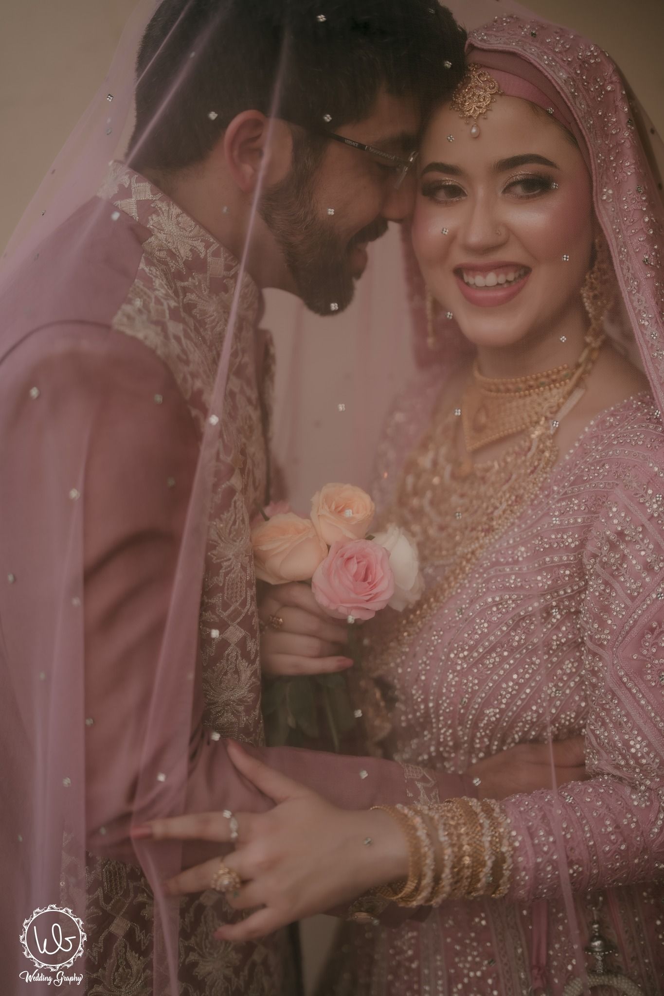 A romantic wedding couple in traditional South Asian attire share intimate moments in soft pink lighting.