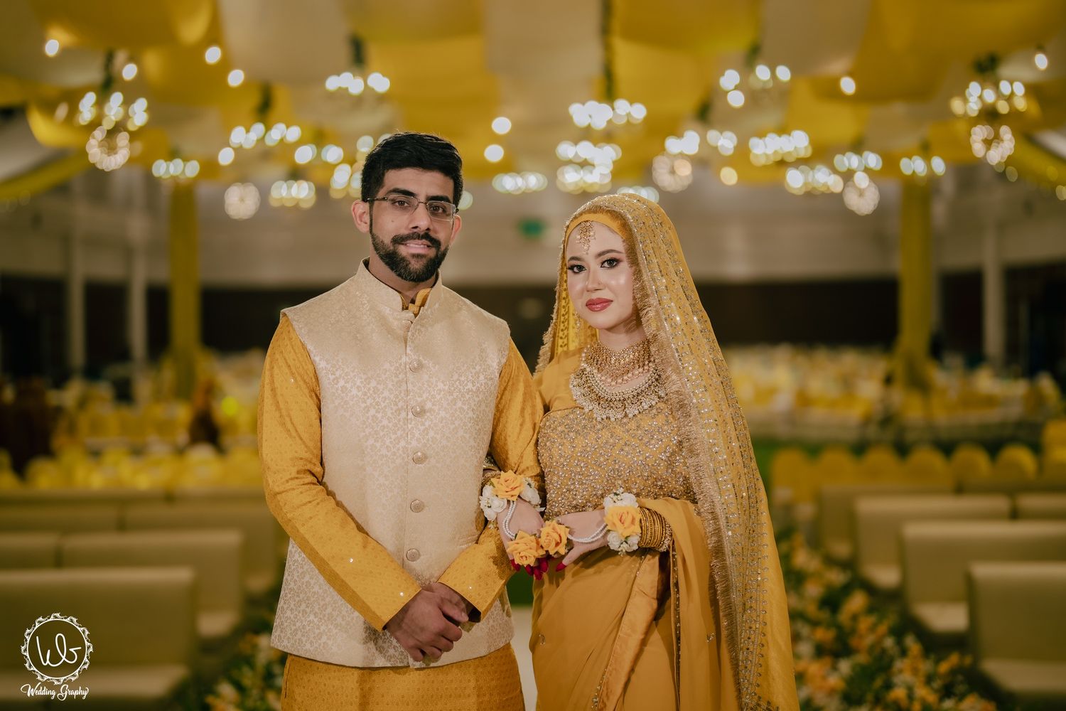 A couple in matching yellow and gold traditional wedding attire standing in an elegantly lit venue.