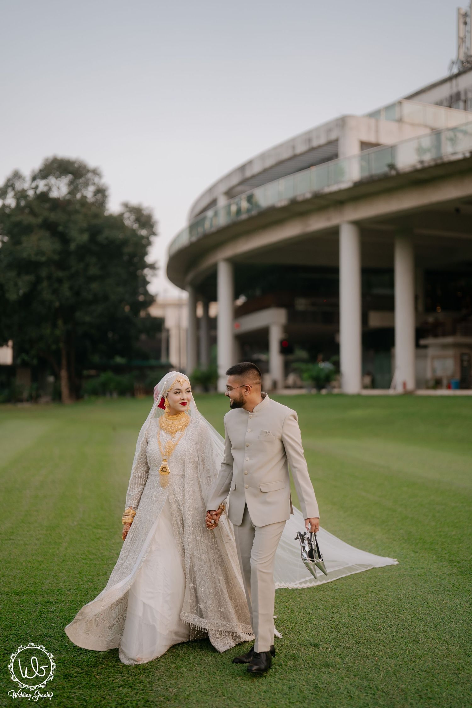 A couple in matching beige traditional outfits walk together on a green lawn beneath an elevated structure.