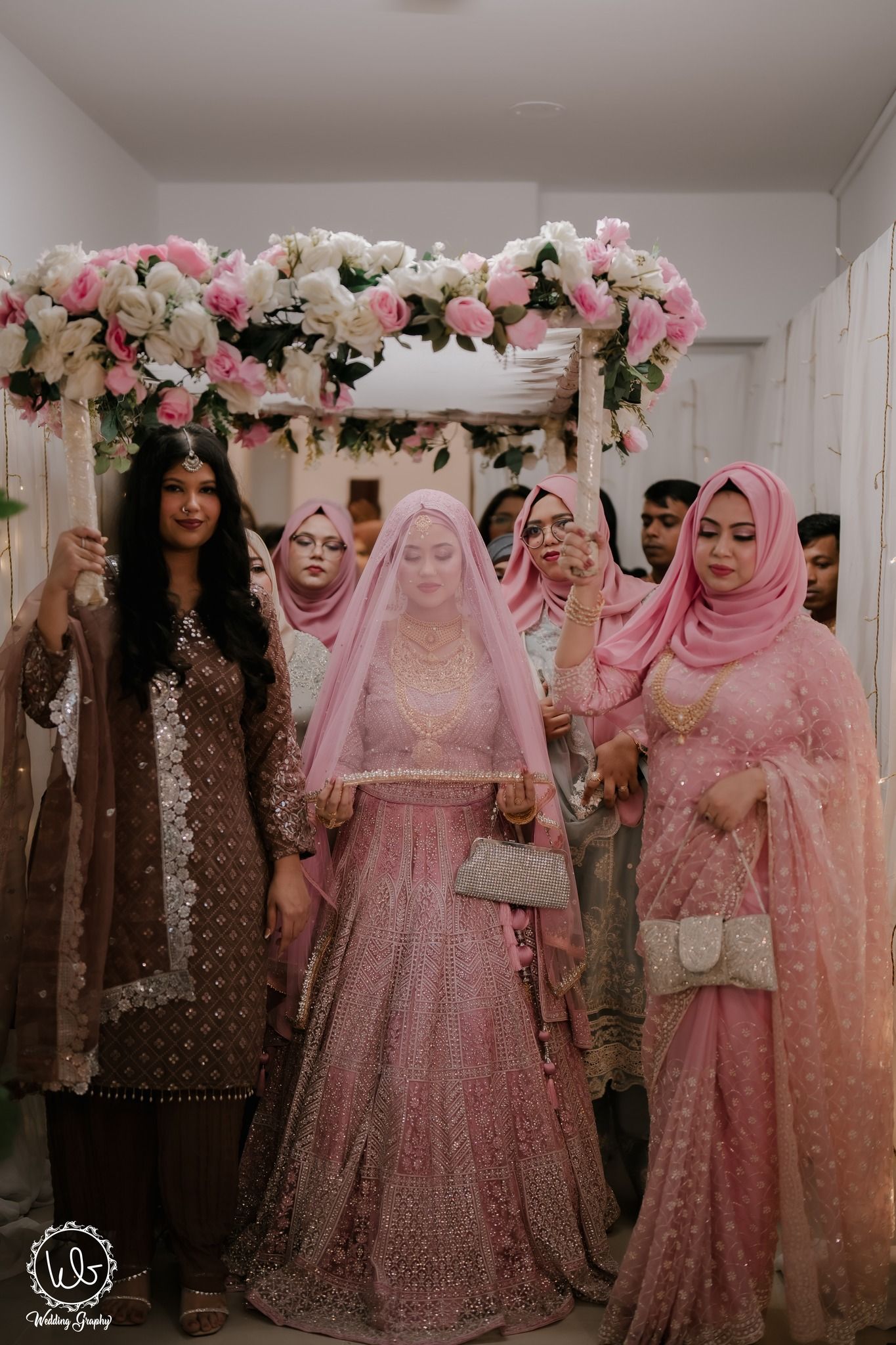 A traditional Muslim wedding ceremony under a floral arch with pink and white roses and attendees in pink attire.