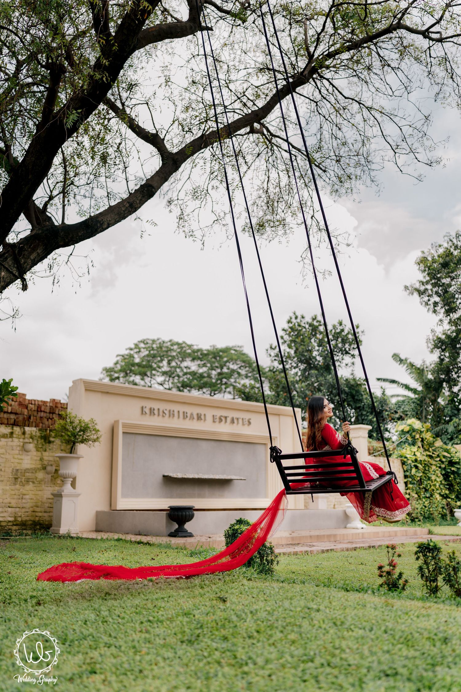 Red saree flowing dramatically on garden swing against cloudy sky.