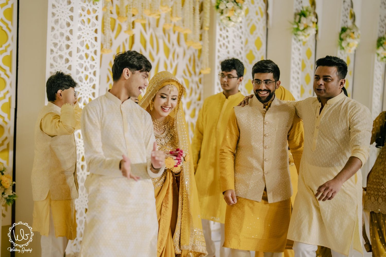 A group celebrates at a festive Indian wedding ceremony decorated in yellow and white floral arrangements.