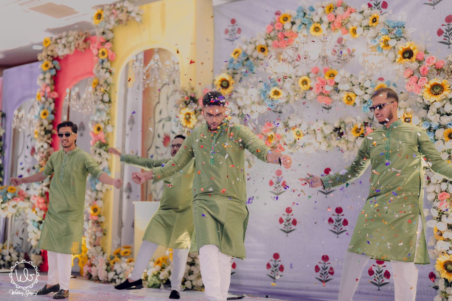 Wedding party dancing against a floral backdrop in coordinated olive green outfits.