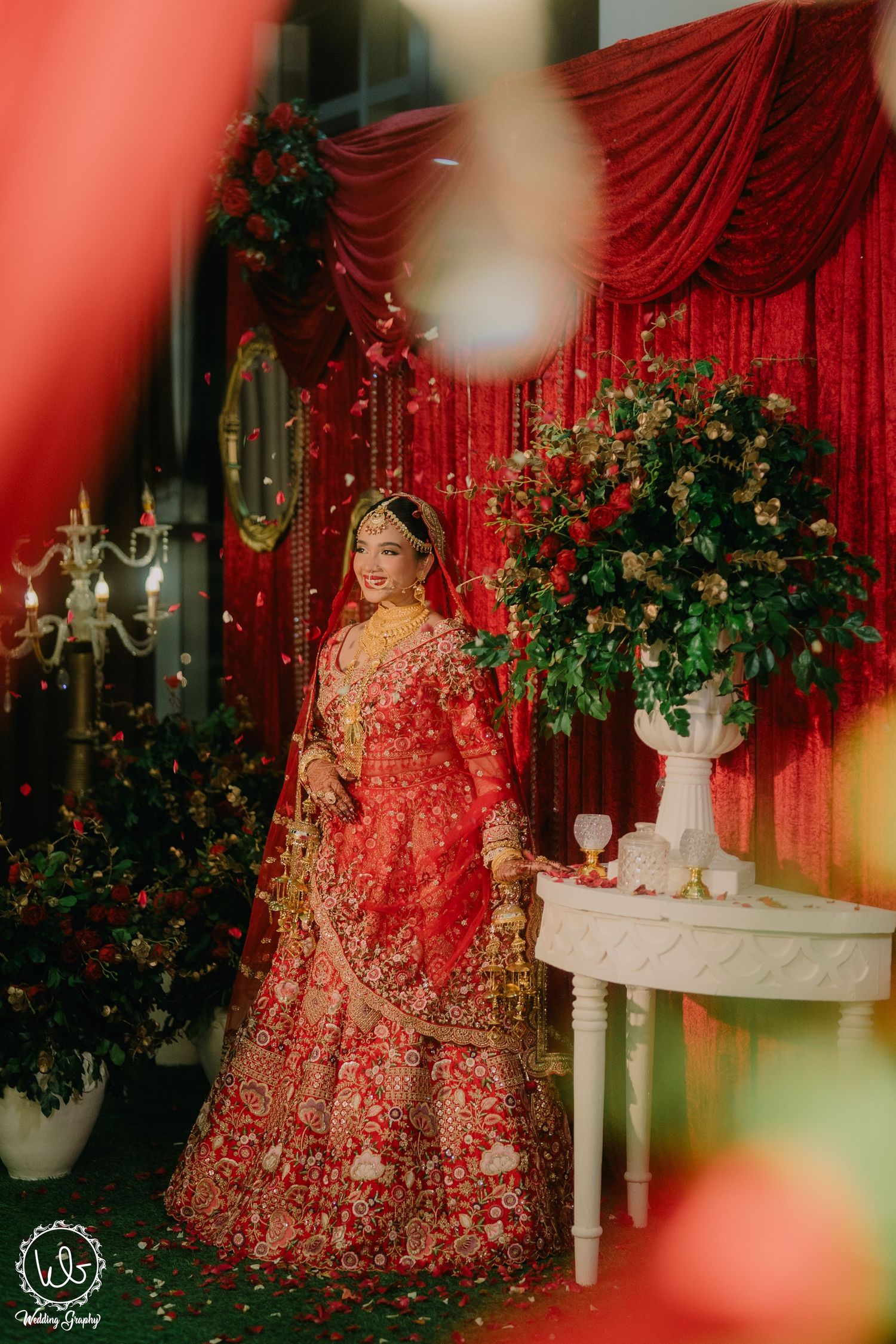 Traditional Indian bride wearing an ornate red lehenga poses by elegant decor with red drapes and floral arrangements.