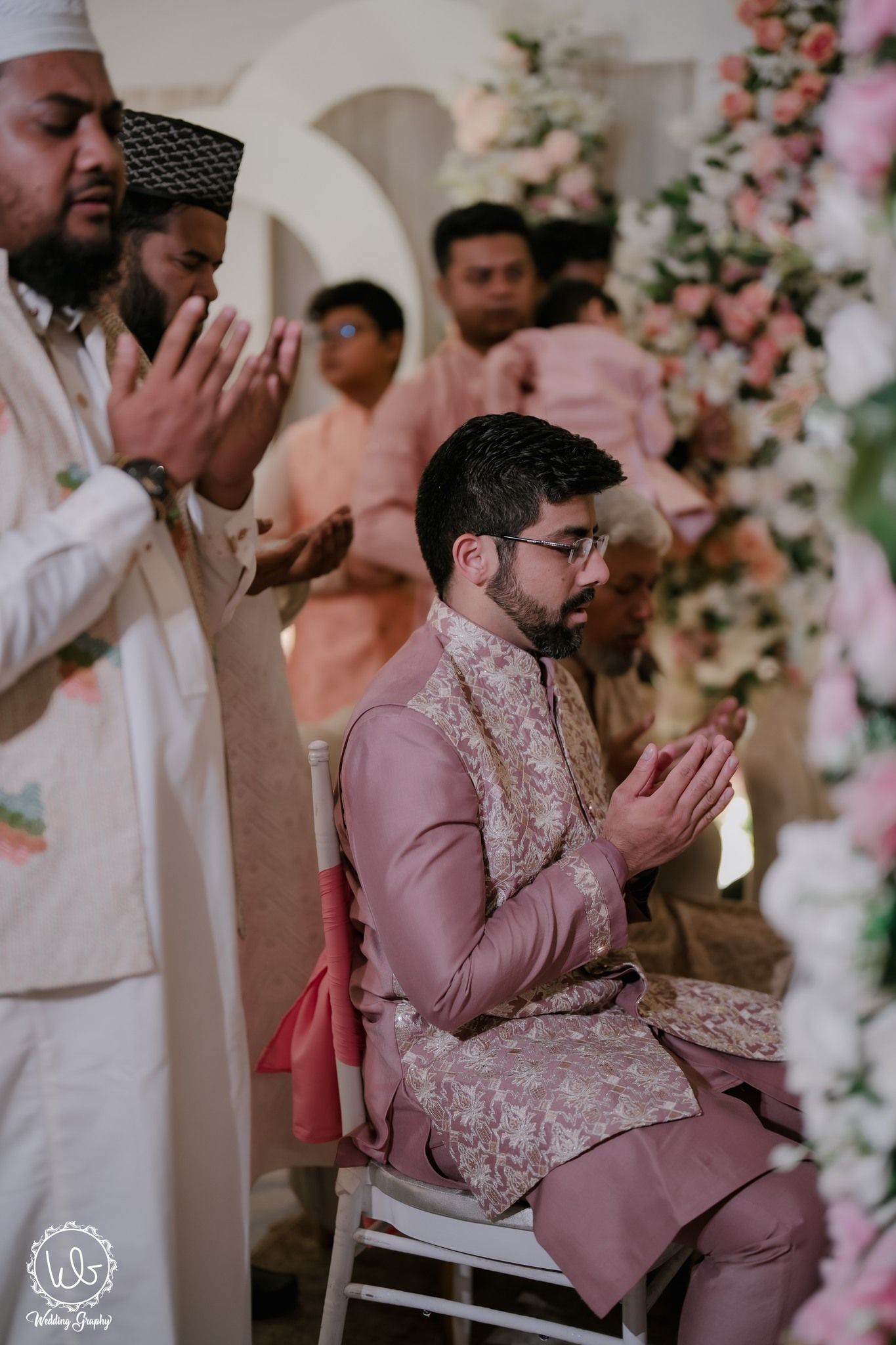 Traditional prayer ceremony with attendees in formal attire seated amidst pink floral decorations.