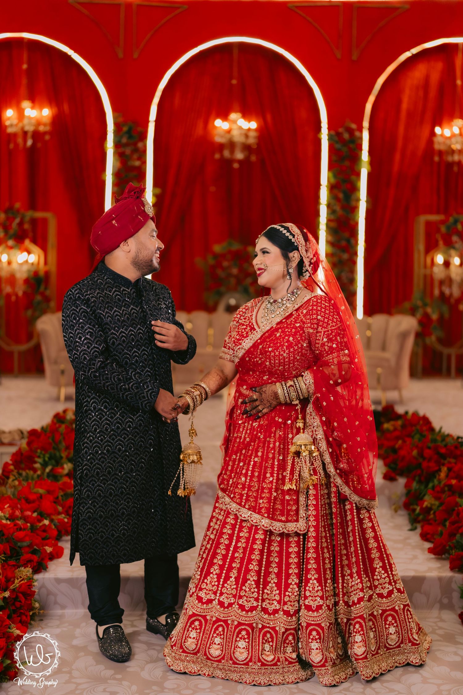 Couple in traditional Indian wedding attire dancing together against a red backdrop with chandeliers.