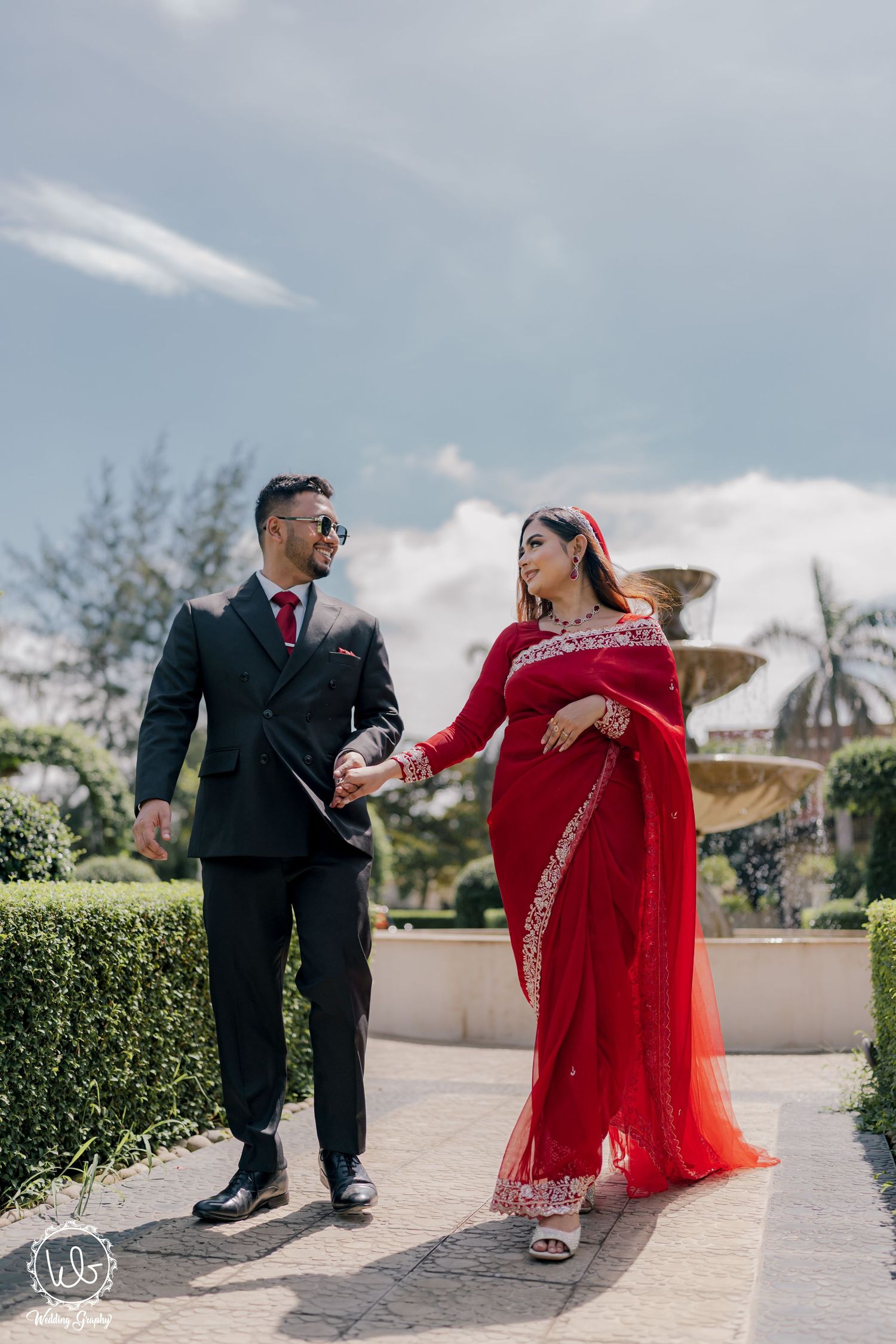 Outdoor wedding photoshoot with couple in red and black attire against sky backdrop.
