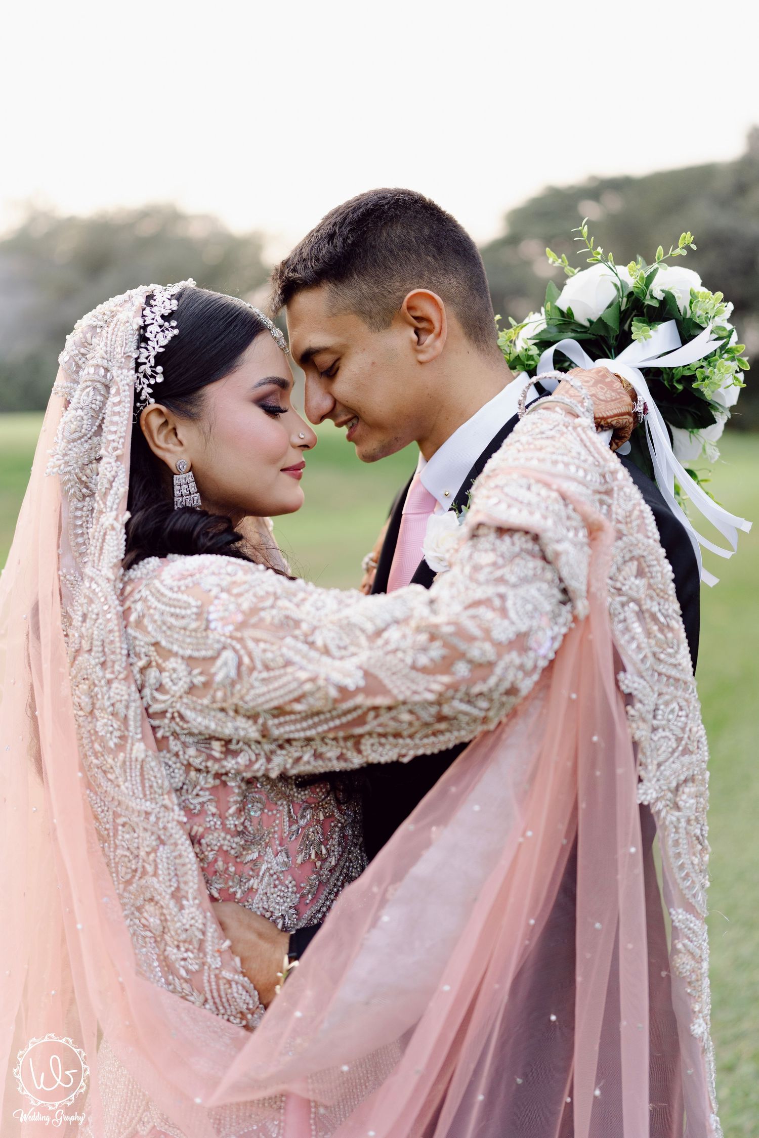 A romantic wedding portrait featuring a couple in traditional South Asian attire with delicate pink veil and white bouquet.