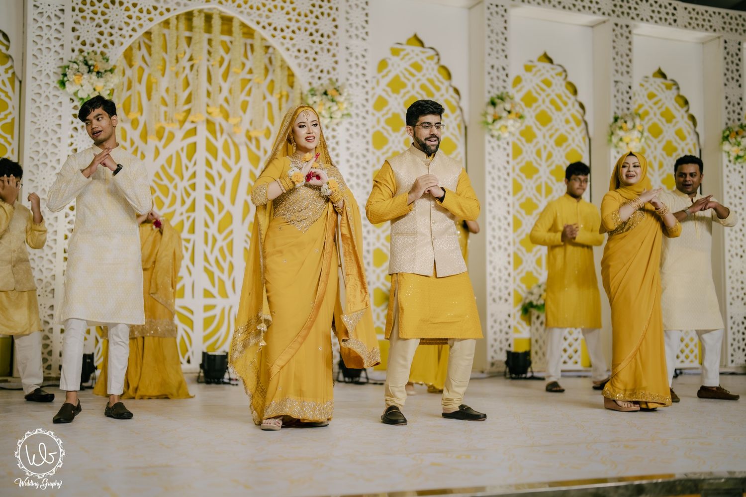 Wedding party members in coordinated yellow and white outfits pose together against an ornate floral backdrop.