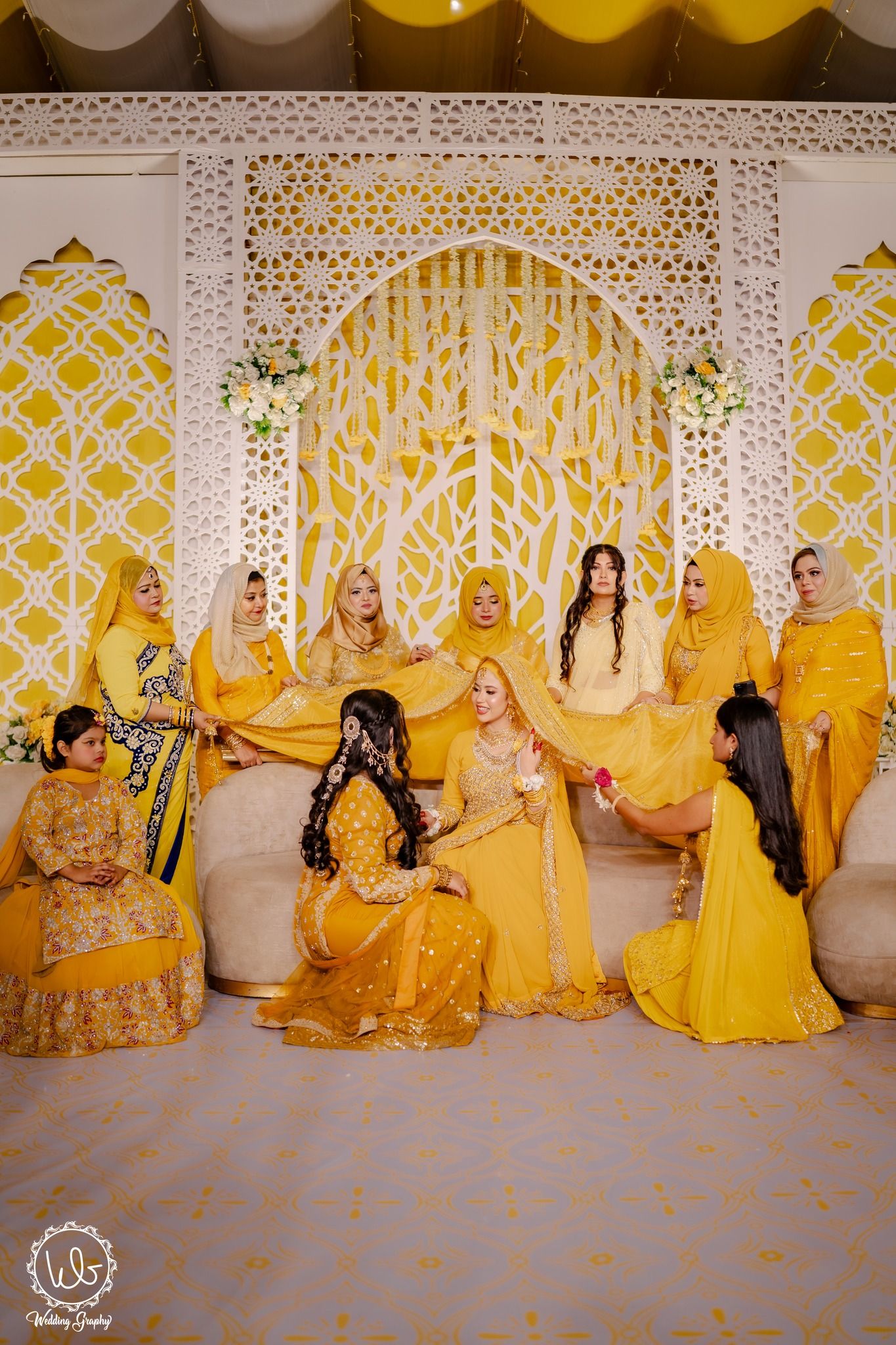 Wedding party in vibrant yellow traditional attire poses against ornate white and gold backdrop at celebration.