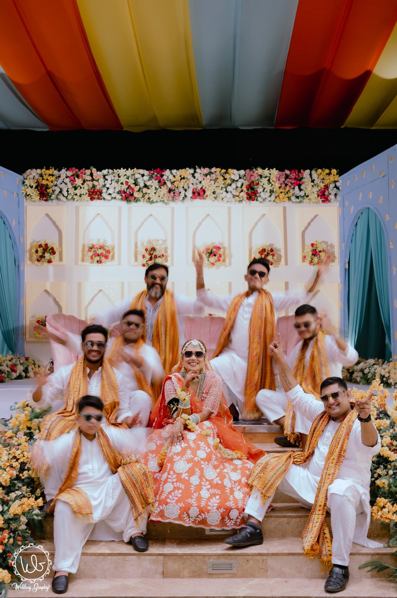 A group in white outfits pose playfully on the floor against a floral backdrop at a formal event.