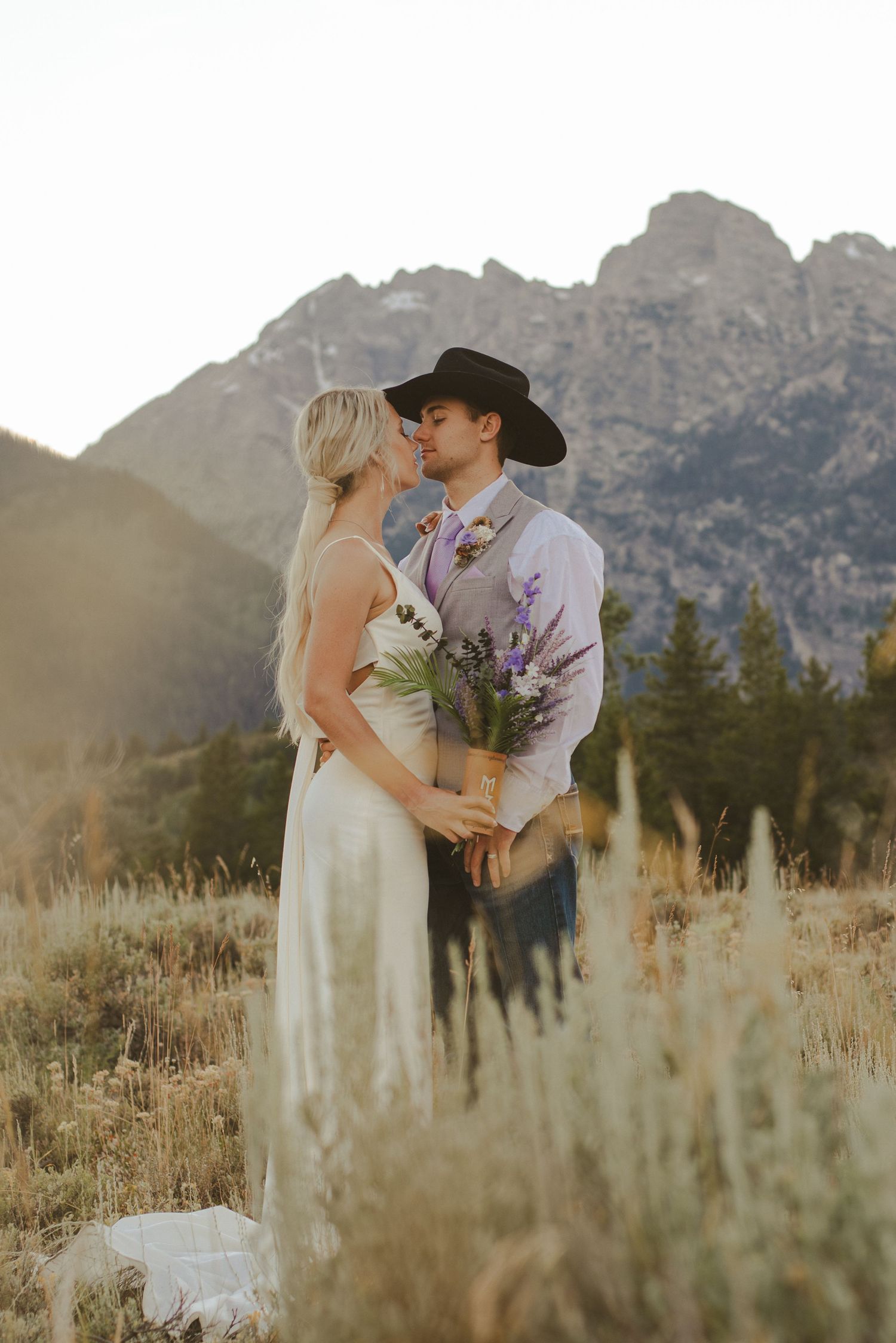 A romantic wedding couple embraces in a field with majestic mountain peaks and golden grass in the background.