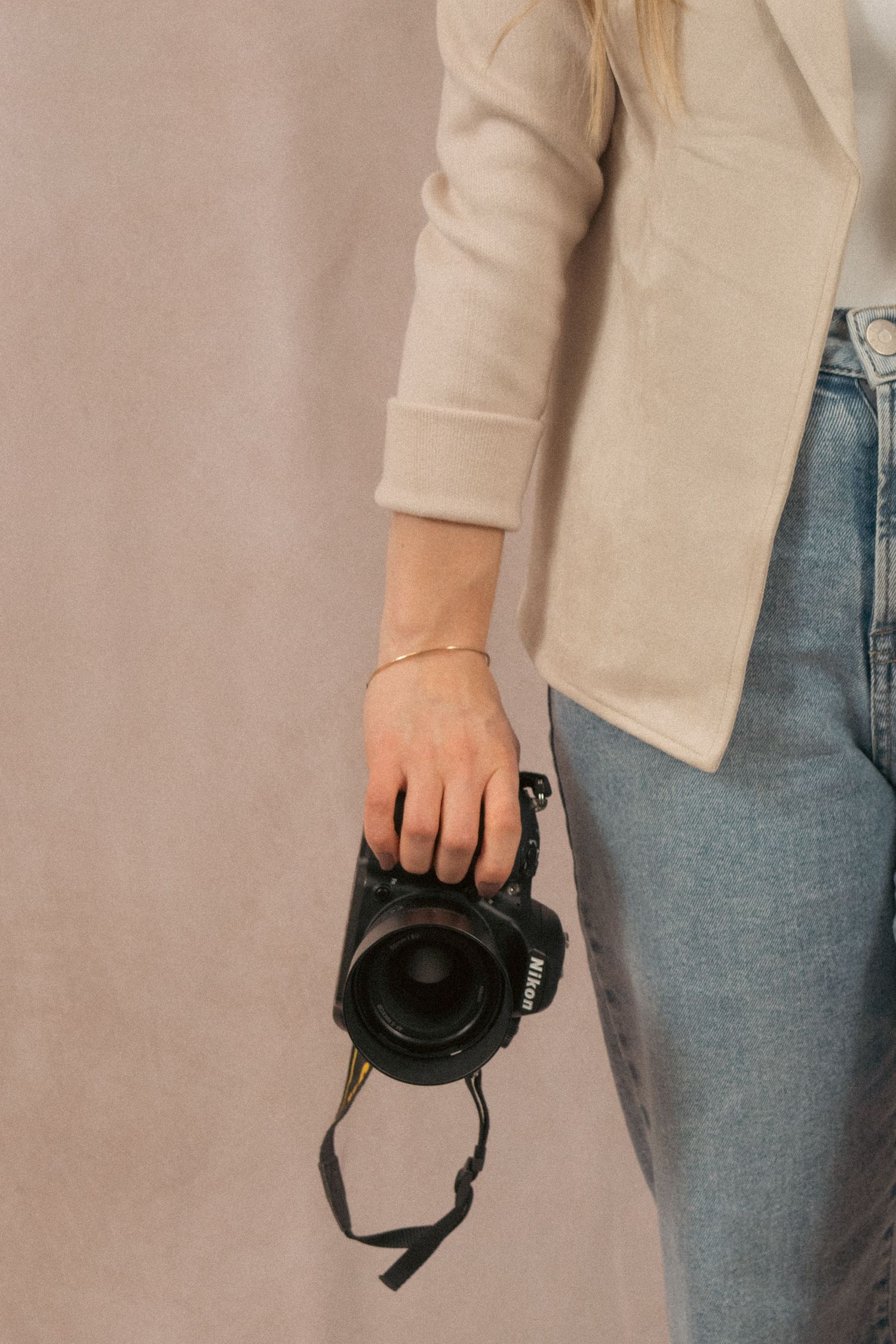 Person wearing cream blazer and jeans holds a black DSLR camera against neutral background.