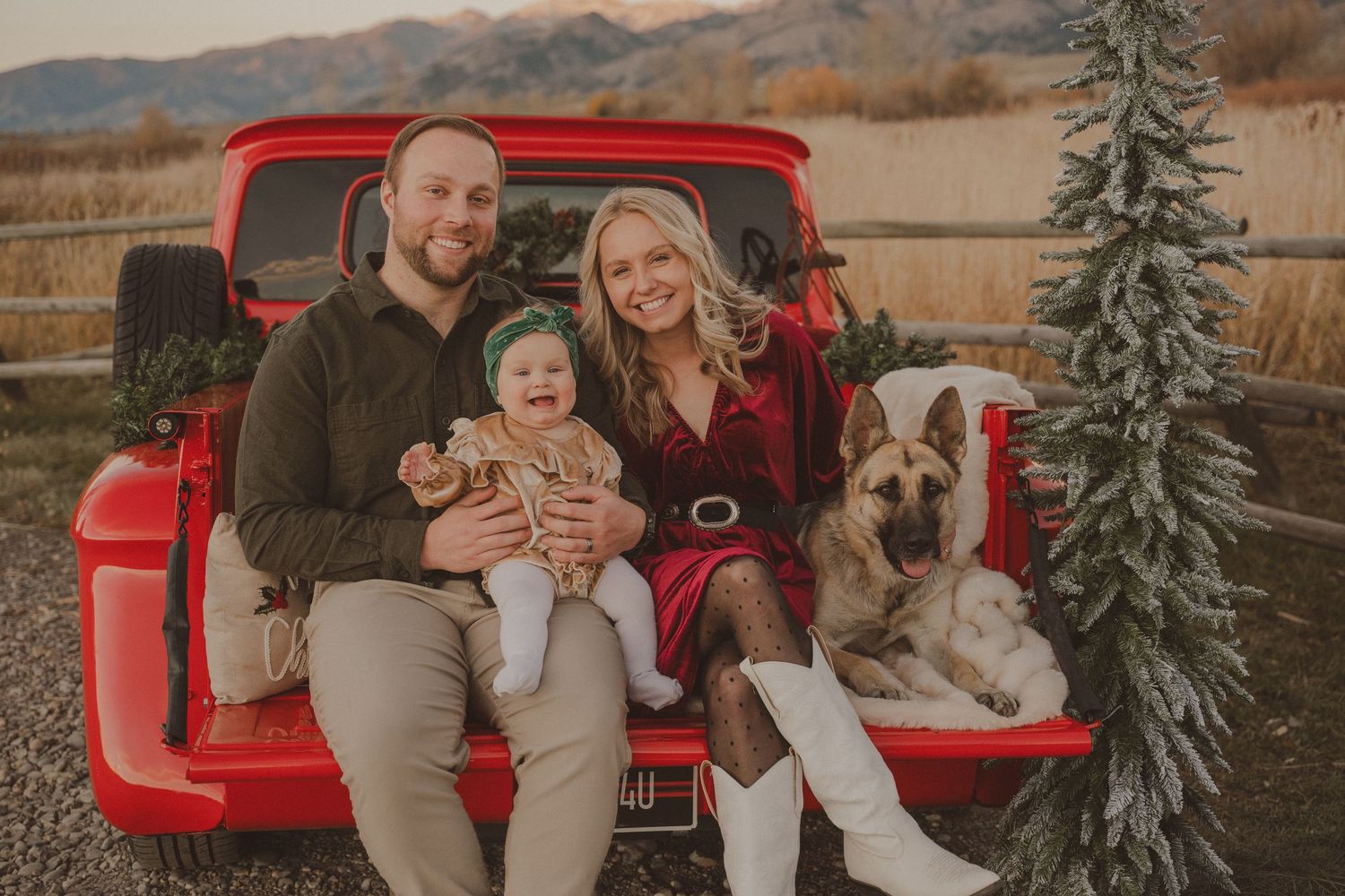 Family poses together on red vintage pickup truck surrounded by evergreen trees in autumn setting.
