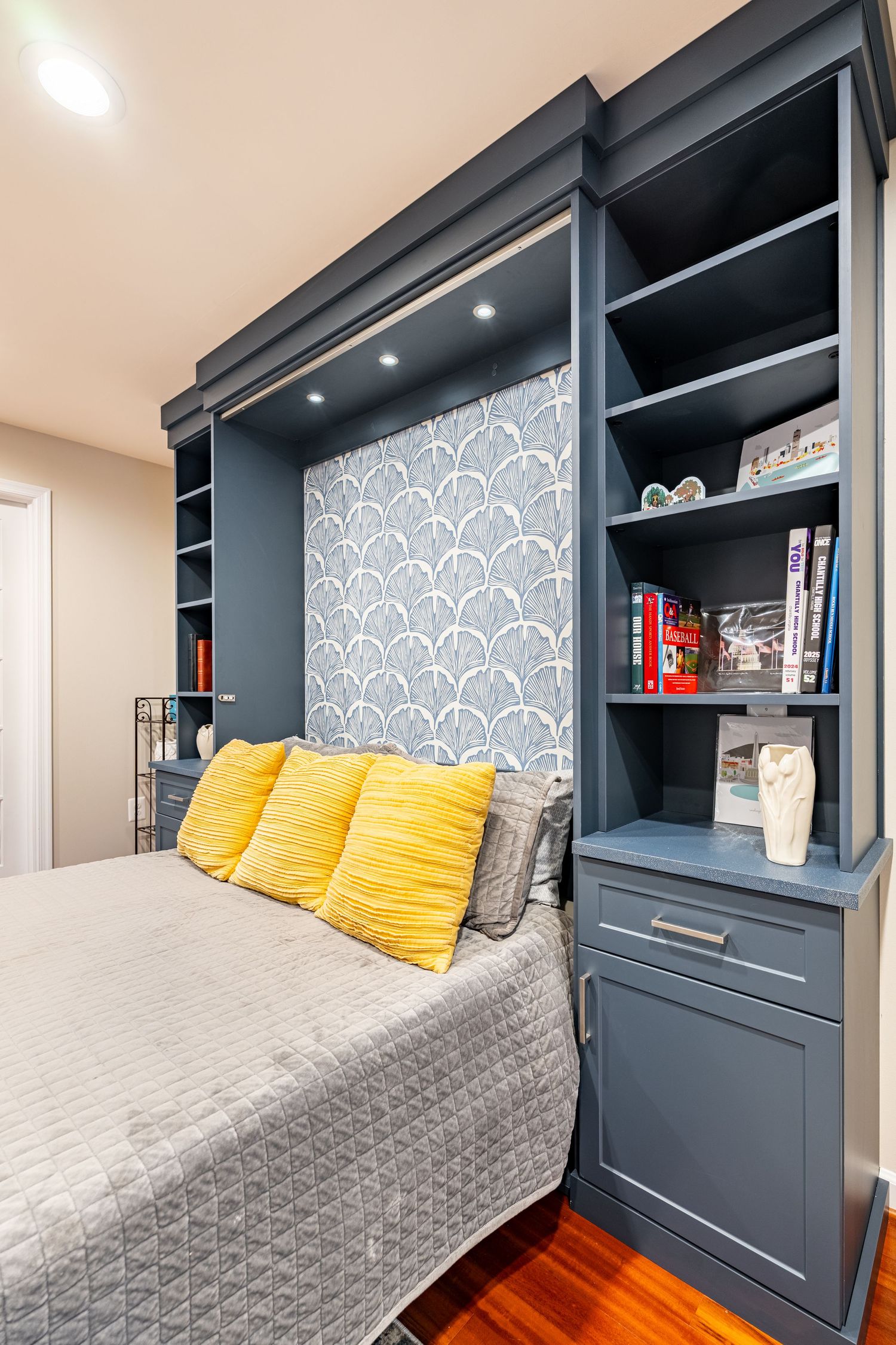 Blue built-in Murphy bed with open shelving, patterned wallpaper headboard, and yellow accent pillows on gray bedding.