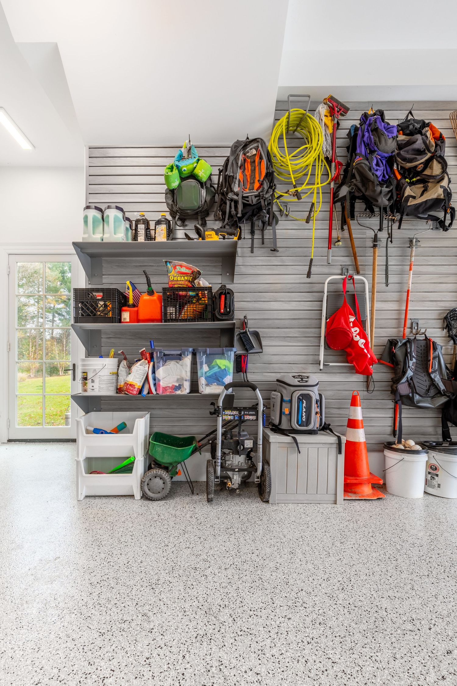 Organized garage wall storage system with slat panels displaying various tools, equipment, and backpacks neatly arranged.