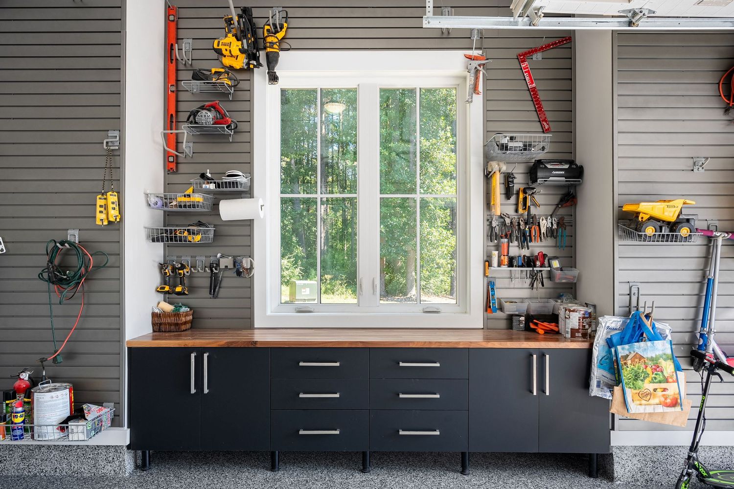 A long gray garage wall with black cabinets, windows, and various tools organized on slatwall storage panels.