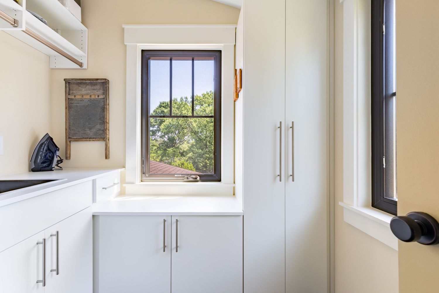 Modern white cabinet storage solutions line the walls of a bright, clean laundry room with natural light from windows.