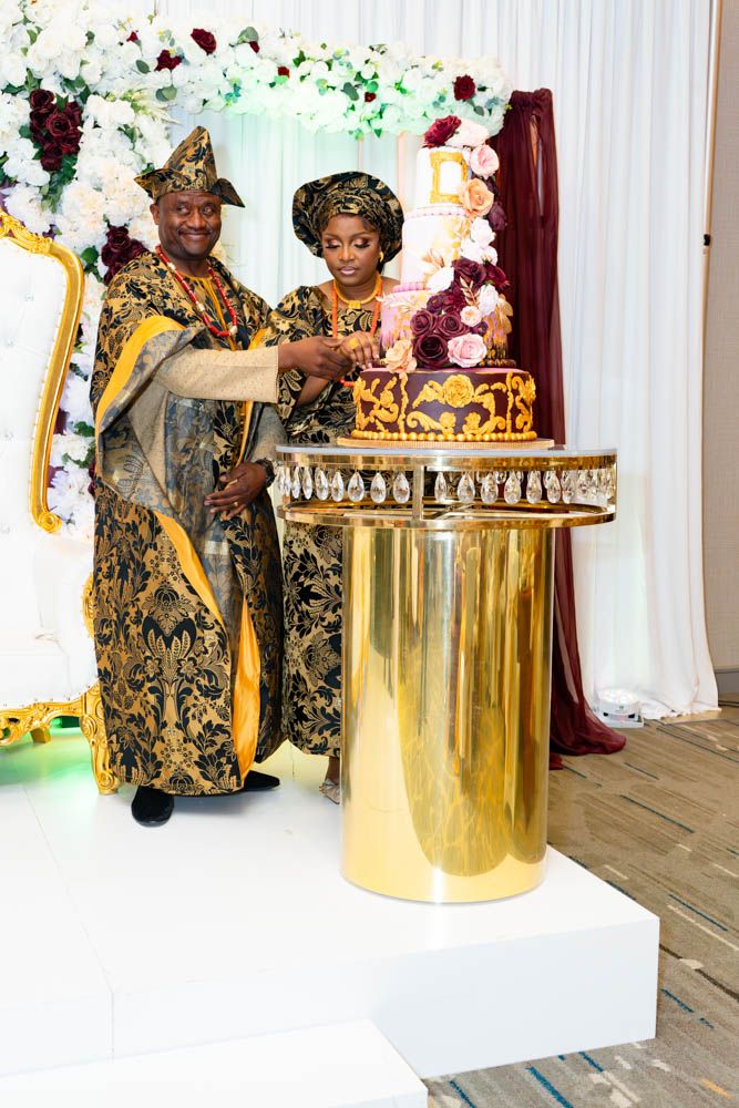 Traditional wedding couple in matching gold and black formal attire beside ornate gold cake display.