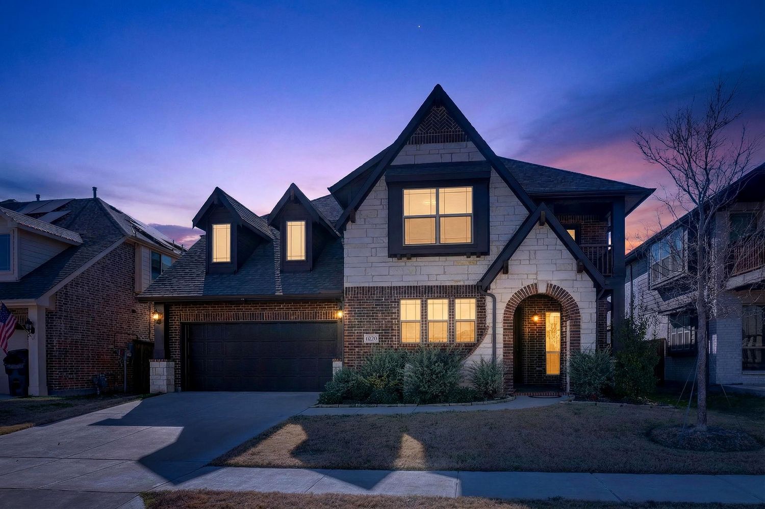 Modern suburban homes with glowing windows illuminate a tranquil dusk scene against a deep blue and purple sky.