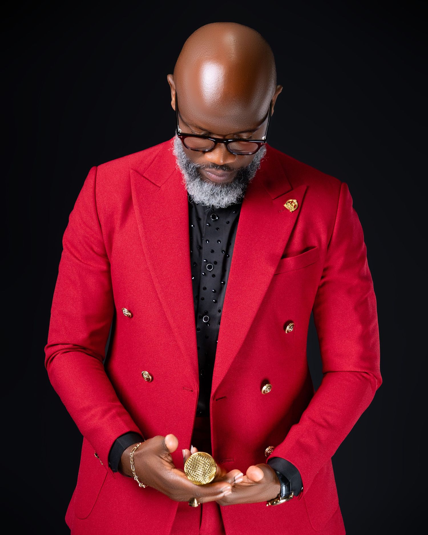 Professional wearing a red blazer and dark turtleneck holds something small in their hands against a black background.