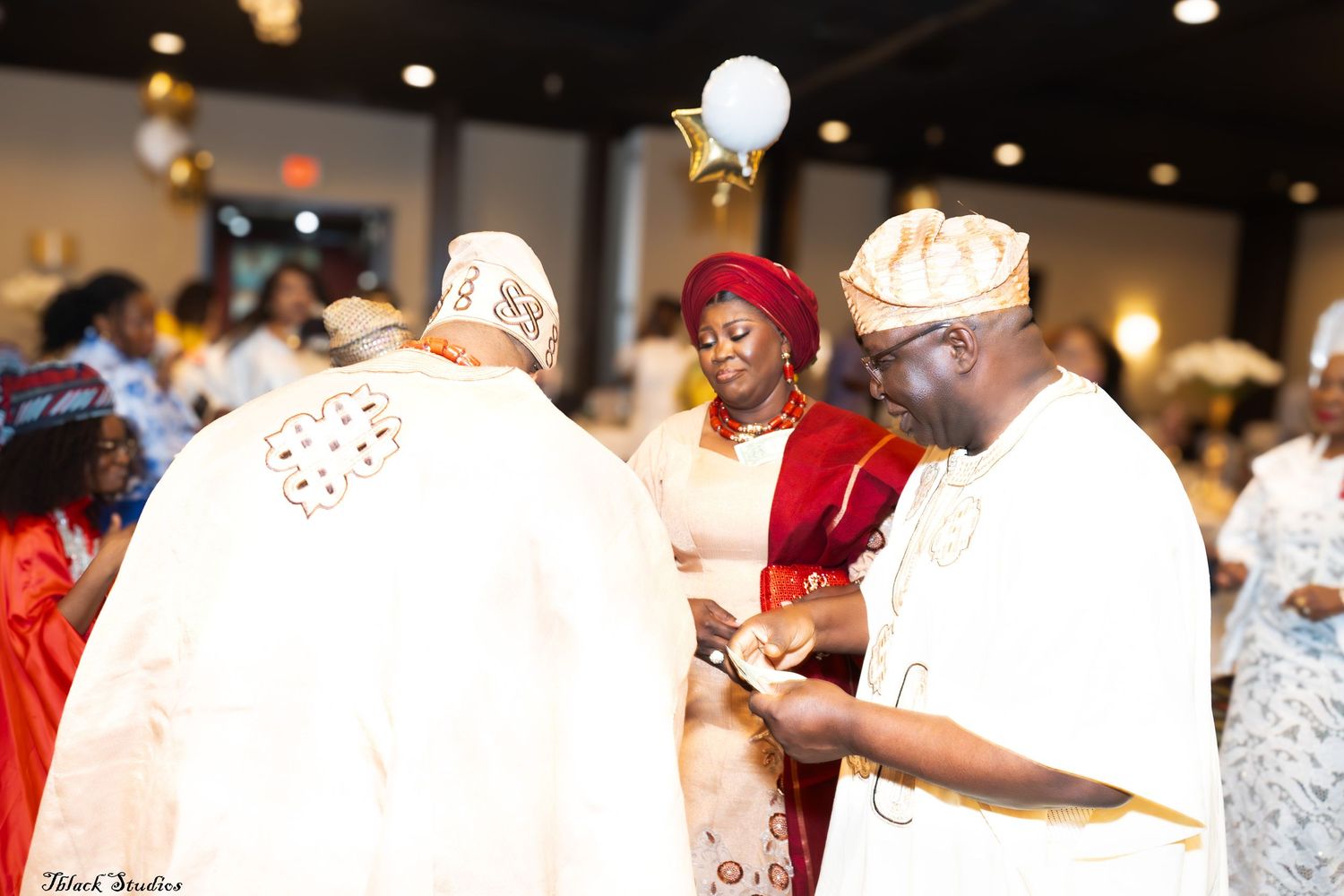 Wedding guests in traditional Nigerian attire celebrating at an indoor reception venue with warm lighting.