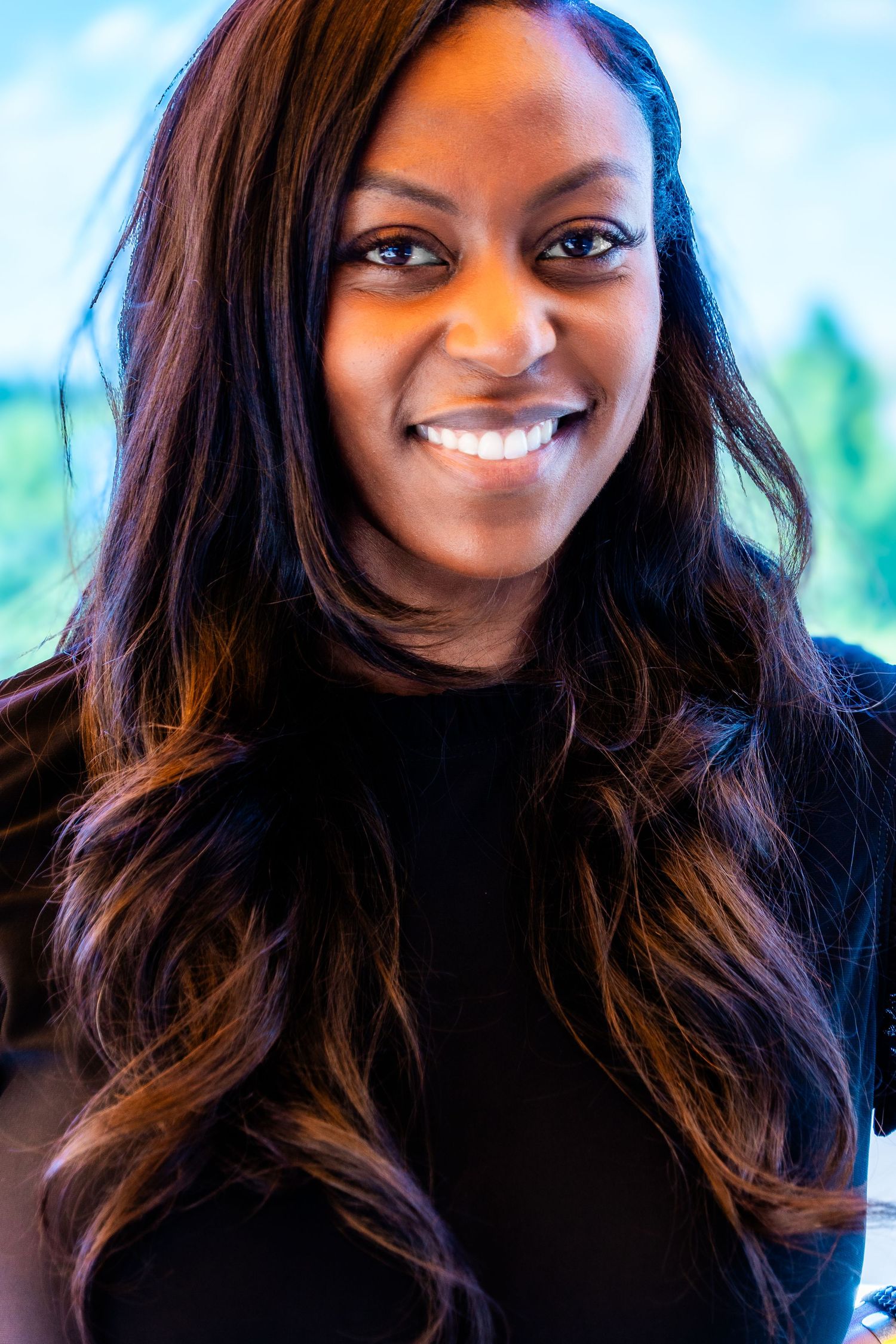 Person in black top with long dark hair smiling against bright background.