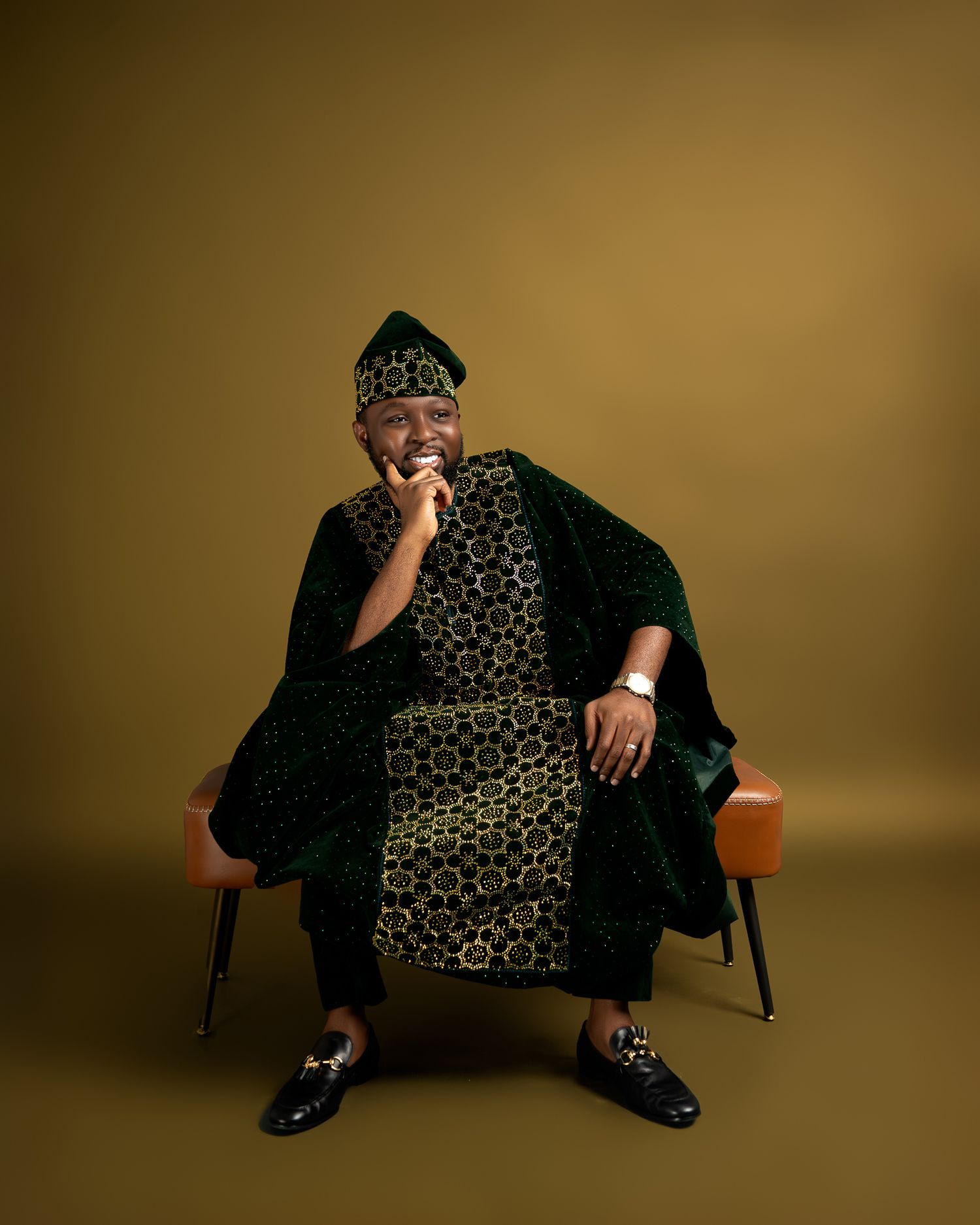 Person in traditional African print attire and hat sits casually in chair against golden backdrop.