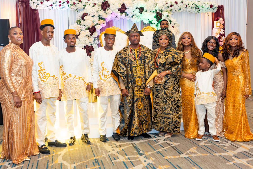 Large group photo of wedding guests in traditional African attire under floral arch decorations.