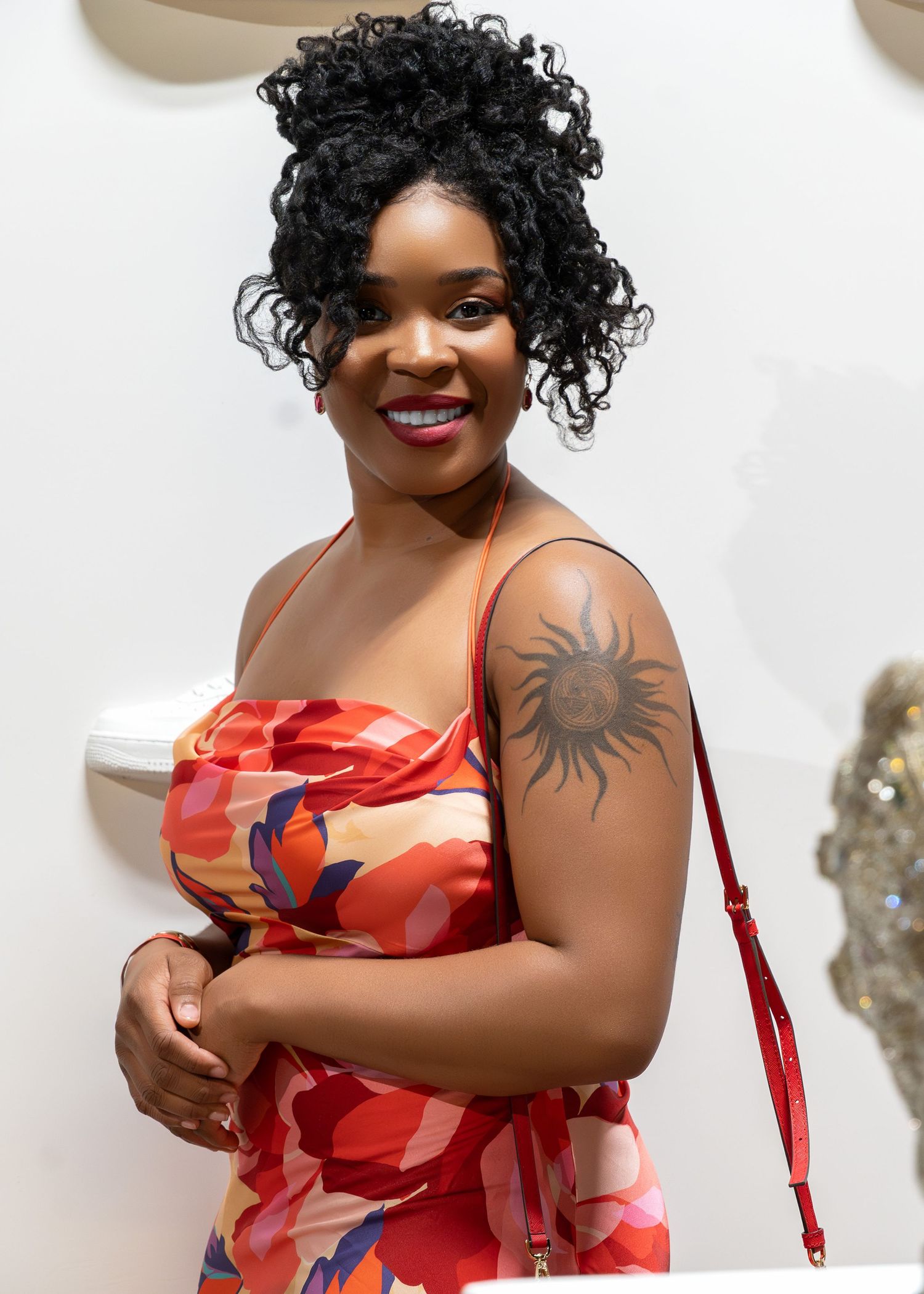 A model in coral floral dress with curly hair poses playfully against white background.