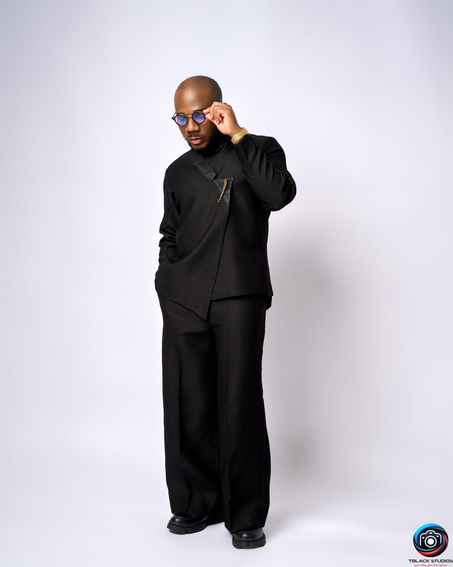 Figure in all-black ensemble poses dramatically against white studio background.