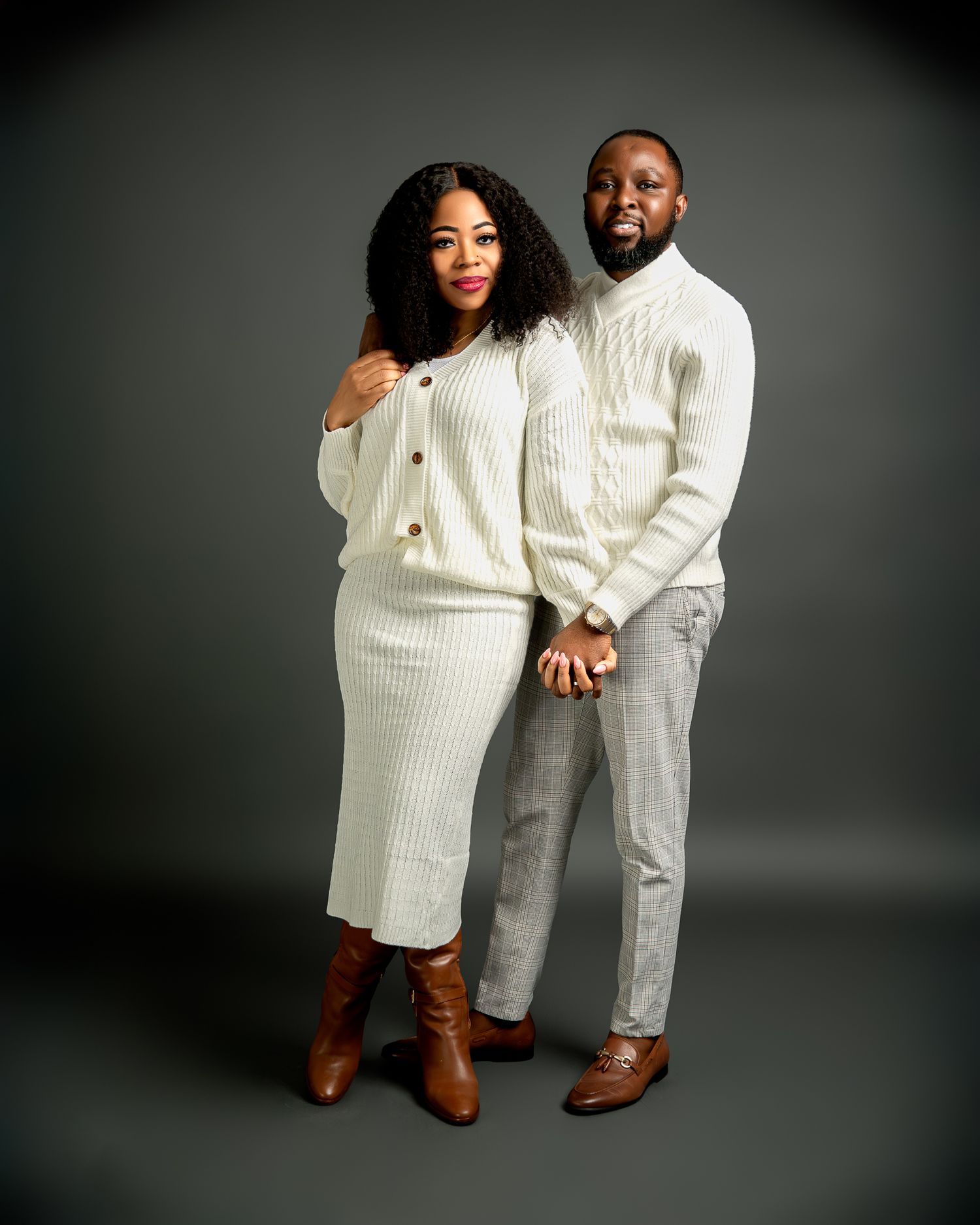 A couple poses together in matching white knit sweaters and brown boots against a dark studio backdrop.