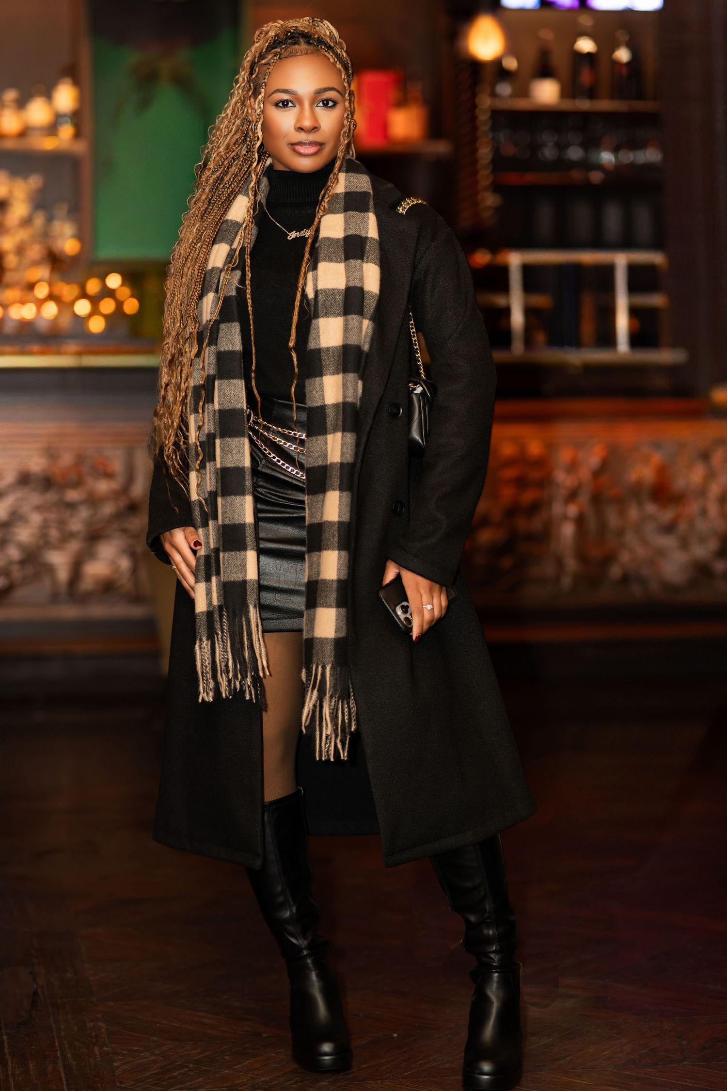 Stylish winter outfit featuring a black coat, plaid scarf, and tall boots in a cozy bar setting with warm lighting.