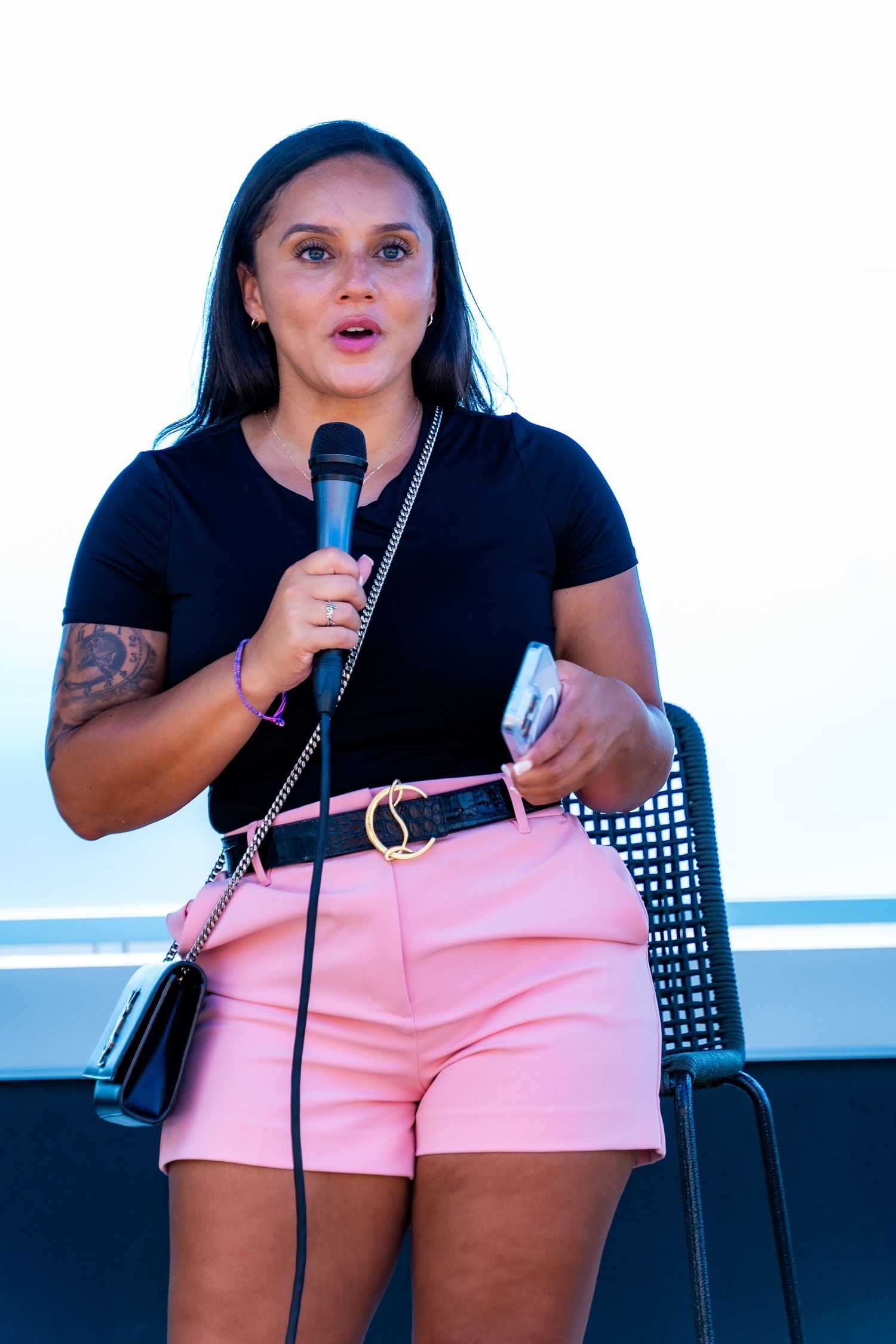 Woman in pink shorts speaking into microphone on outdoor stage.