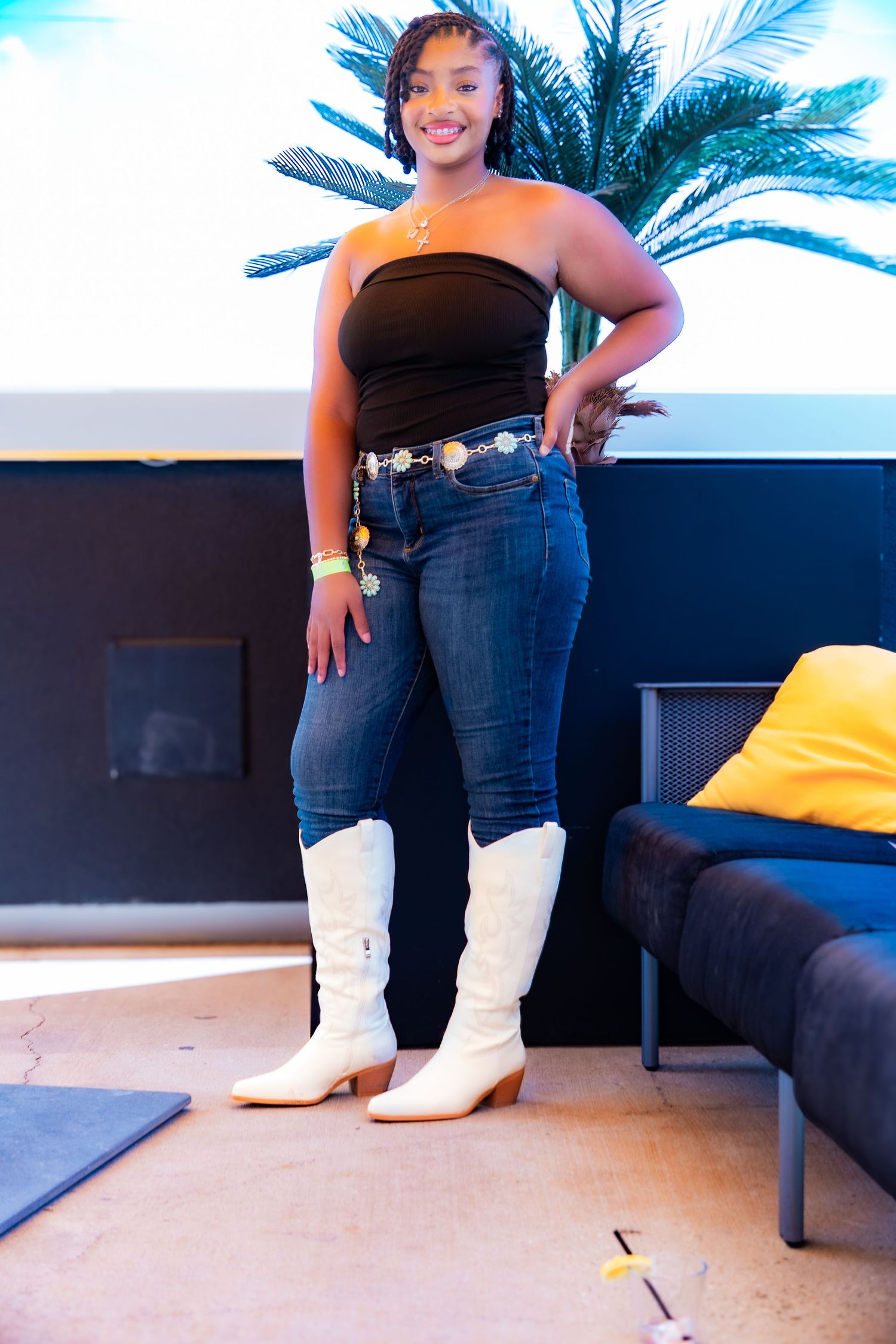 Woman wearing black top, jeans and white boots poses near palm trees.