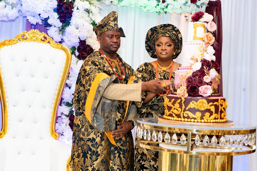 Couple in ornate gold and black traditional attire cutting an elegant tiered wedding cake.