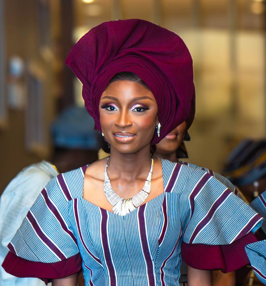 Elegant wedding attendant wearing a burgundy gele headwrap and blue striped traditional dress with statement necklace.